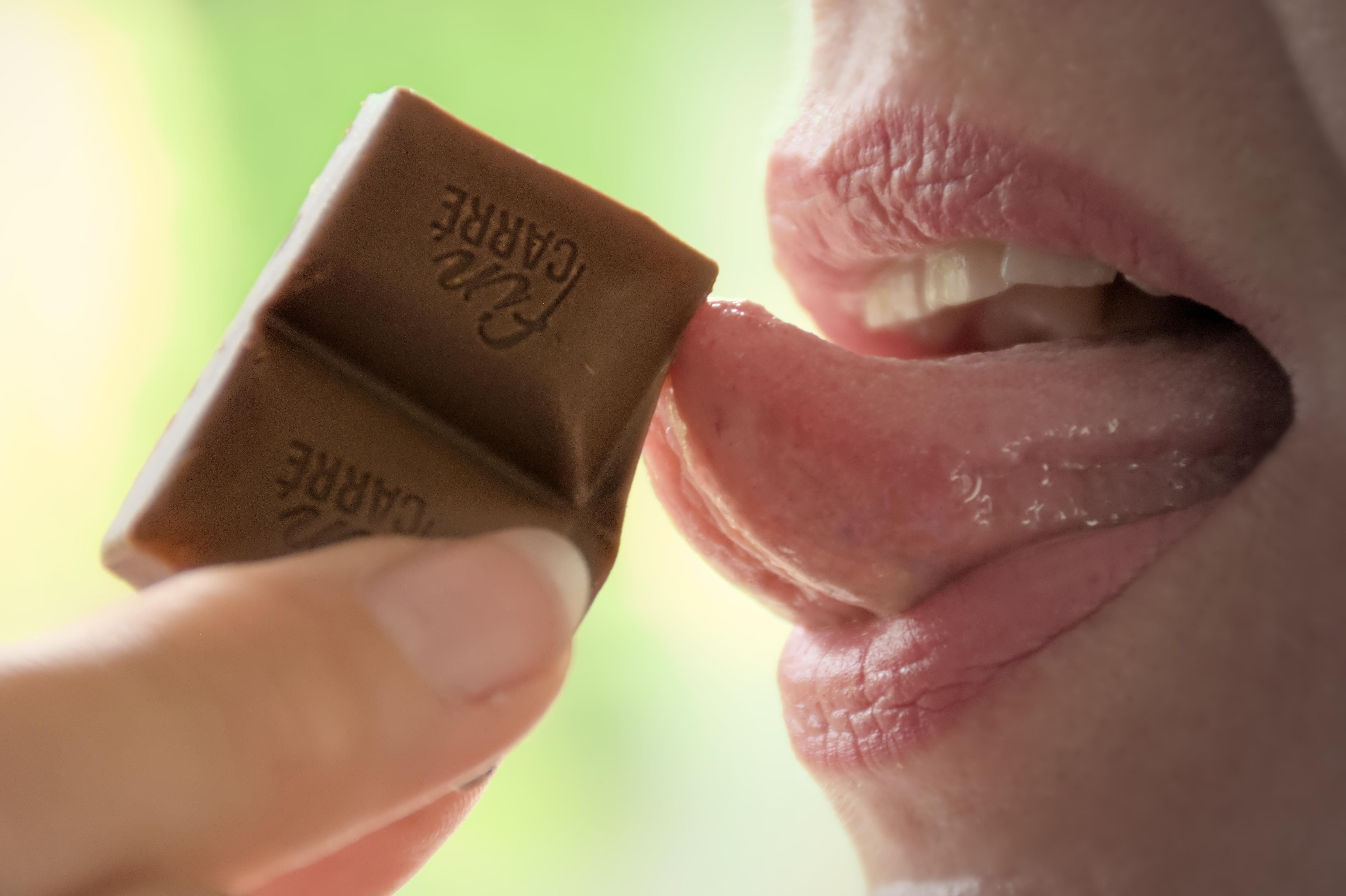 Close-up of a person holding a piece of chocolate near their mouth with the tongue extended to touch it. The chocolate is embossed with text. Lips are parted, teeth slightly visible, and a neutral background is present.
### 
Nahaufnahme einer Person, die ein Stück Schokolade in der Nähe ihres Mundes hält und die Zunge ausstreckt, um es zu berühren. Die Schokolade ist mit Text geprägt. Die Lippen sind gescheitelt, die Zähne sind leicht sichtbar und es ist ein neutraler Hintergrund zu sehen.