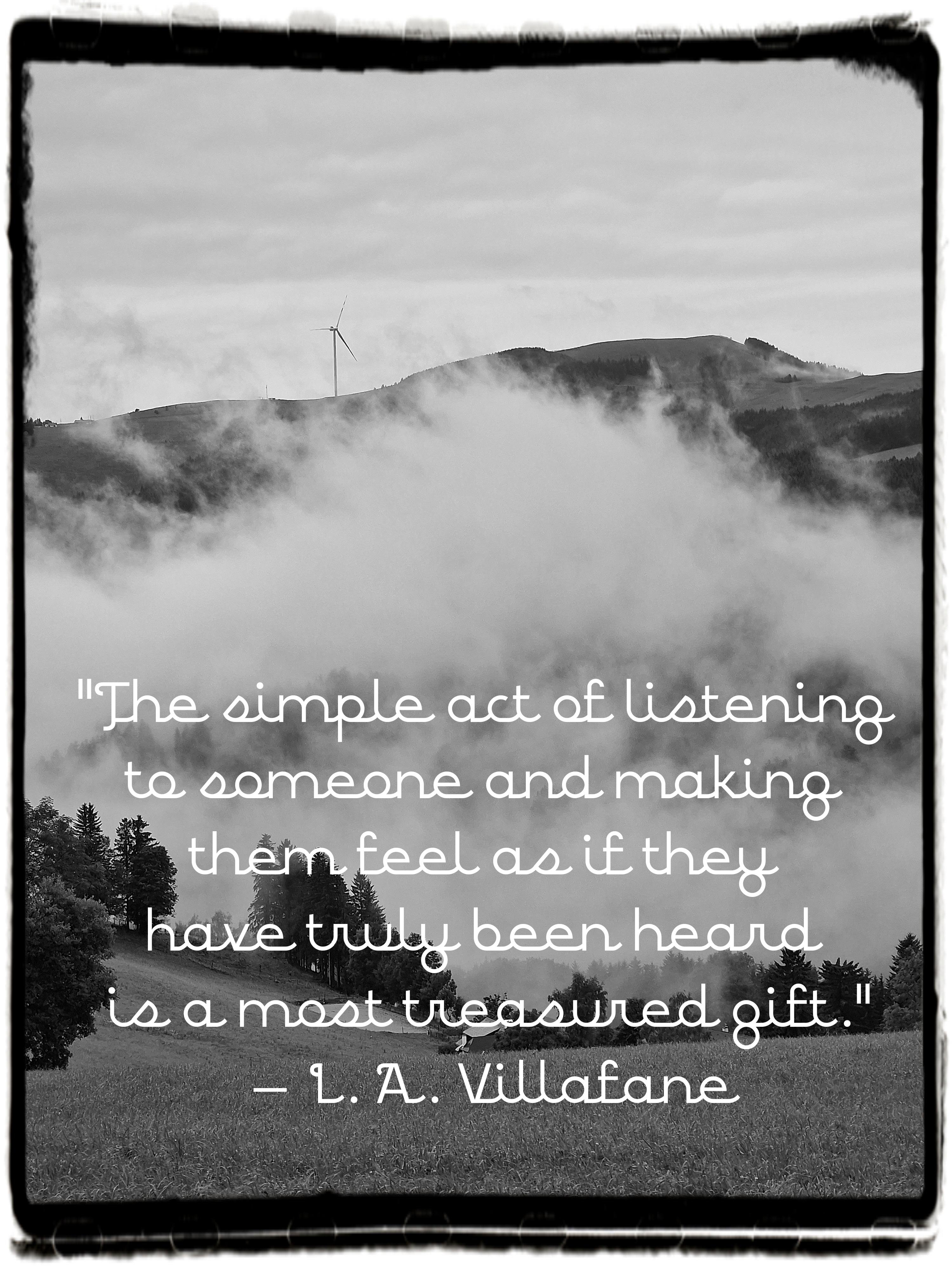 Black-and-white landscape photo framed with a rough, film-strip-style border. The scene shows rolling hills partly hidden beneath thick, low-hanging clouds that drift across the middle of the image. A single wind turbine stands on a distant ridge, barely visible through the mist. The foreground features a sloping meadow with scattered dark evergreen trees, creating a calm contrast with the soft, enveloping fog. Near the bottom, centered over the landscape, a white cursive font displays the quote: "The simple act of listening to someone and making them feel as if they have truly been heard is a most treasured gift." – L. A. Villafane. The overall mood is serene, reflective, and quietly emotional.