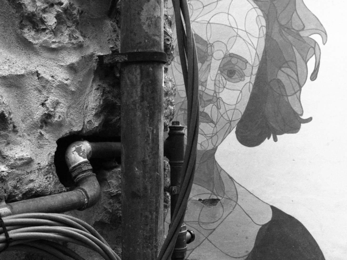 Mural di una donna stilizzata, nei colori della terra, che sbuca, come a sbirciare, da un angolo di muro ingombro di tubi e cavi. Foto in bianco e nero.
