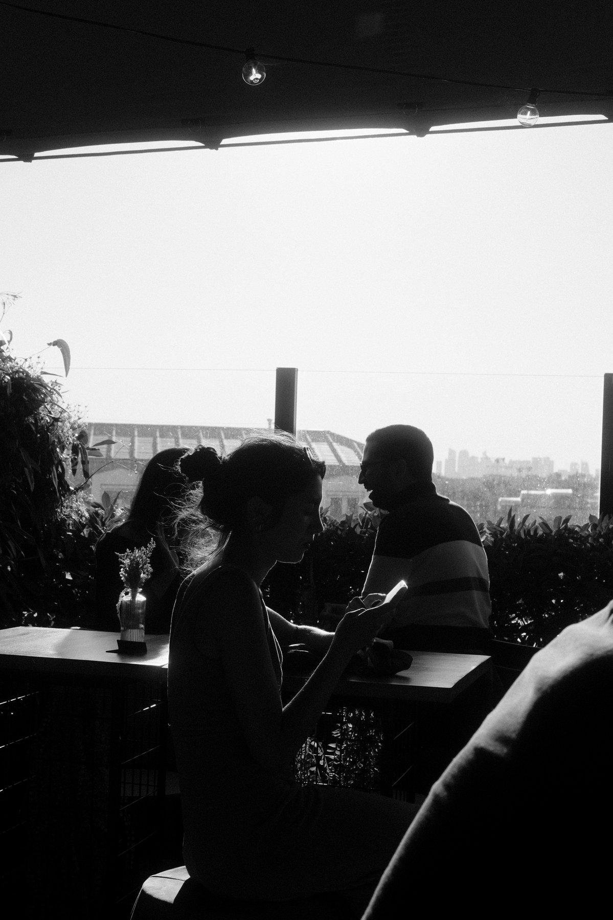 Photo verticale. Silhouette de profile du visage d’une jeune femme avec les cheveux en chignon, en train de consulter son téléphone. Dans l’arrière plan, un homme de profil dans l’autre sens rigole en face de quelqu’un à moitié caché par la jeune femme