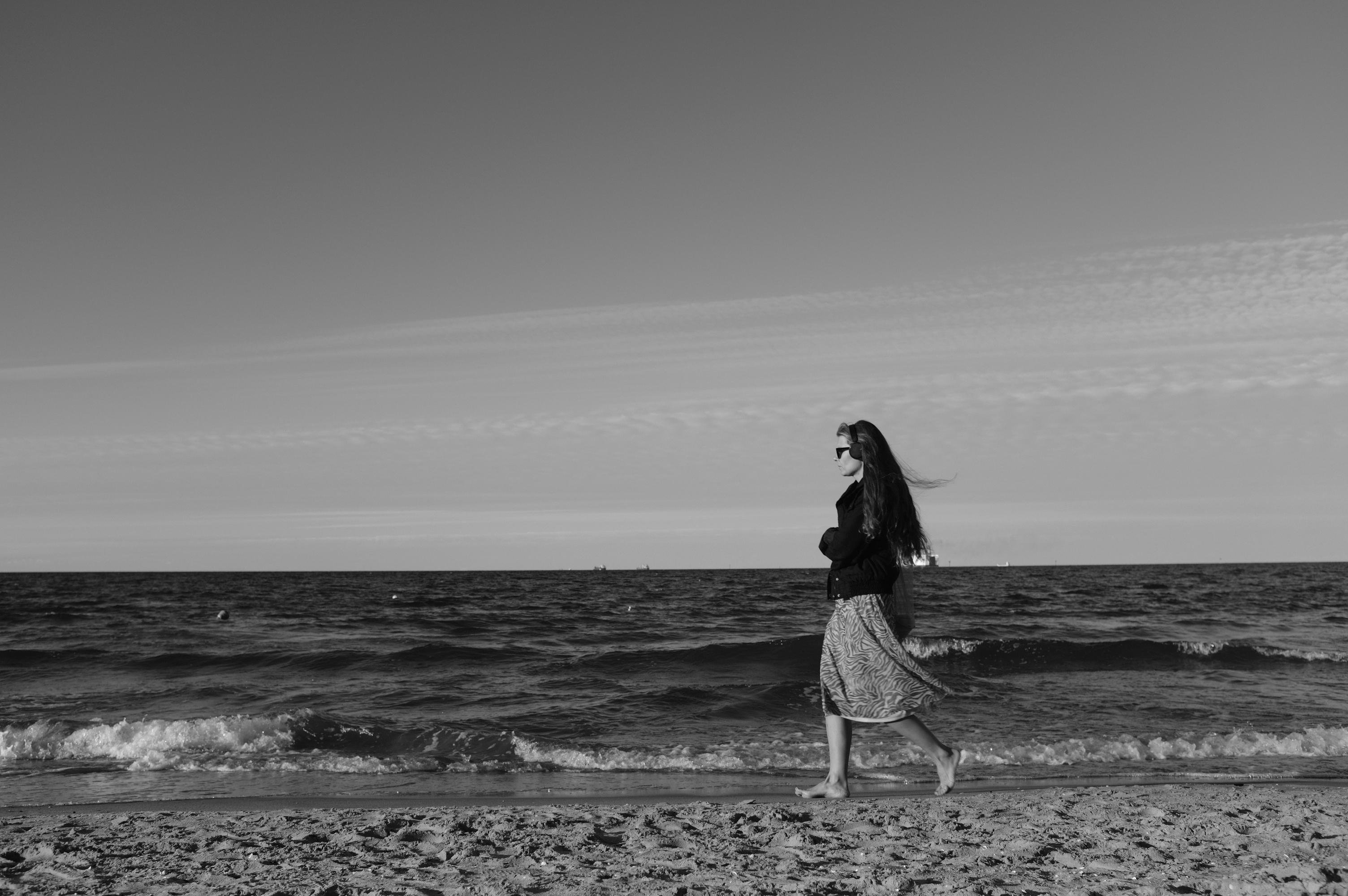 A woman with long hair walks barefoot along a sandy beach, wearing a long skirt and jacket. The ocean waves appear to be calm, and the sky above is clear with streaks of clouds. The image is in black and white, creating a serene atmosphere.