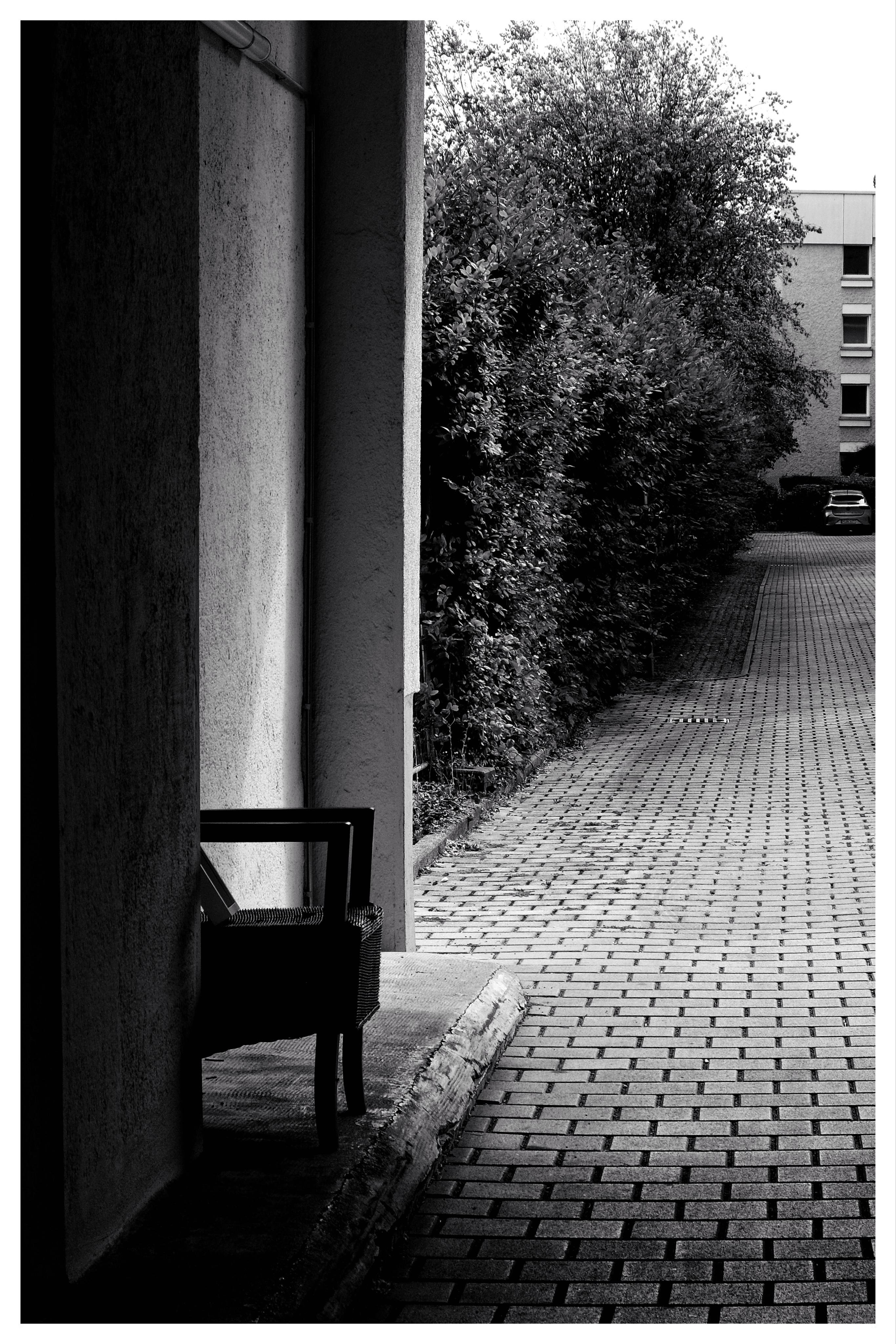 Black-and-white photo of a brick-paved walkway beside a building wall; a single chair sits in shadow under a covered entry on the left, dense bushes line the right side, and an apartment building and parked car appear in the distance.