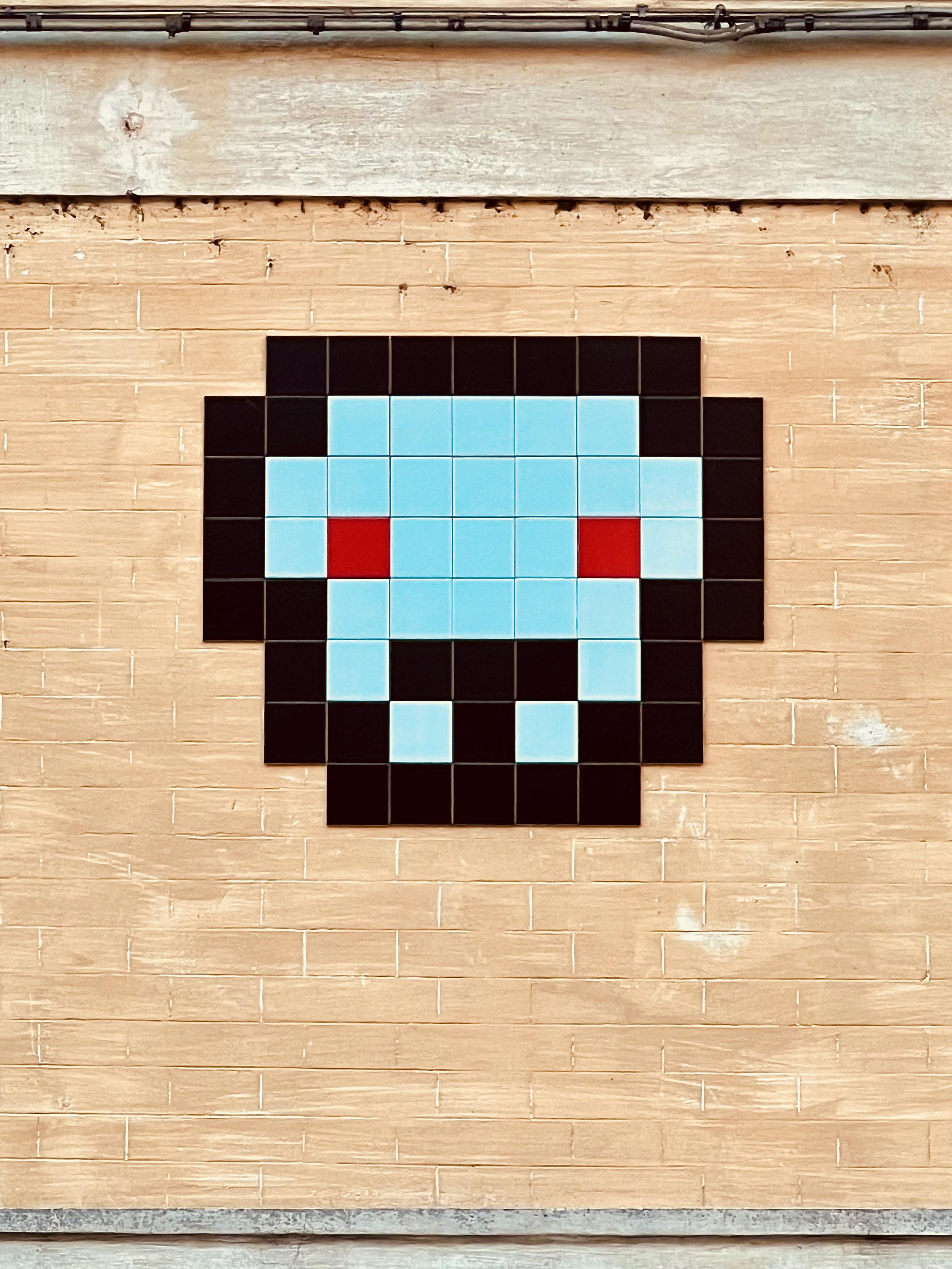 Mosaico in pixel art su un muro, che raffigura un personaggio o volto stilizzato. È composto da tessere nere, blu e rosse disposte in un motivo. Lo sfondo è un muro di mattoni beige.
