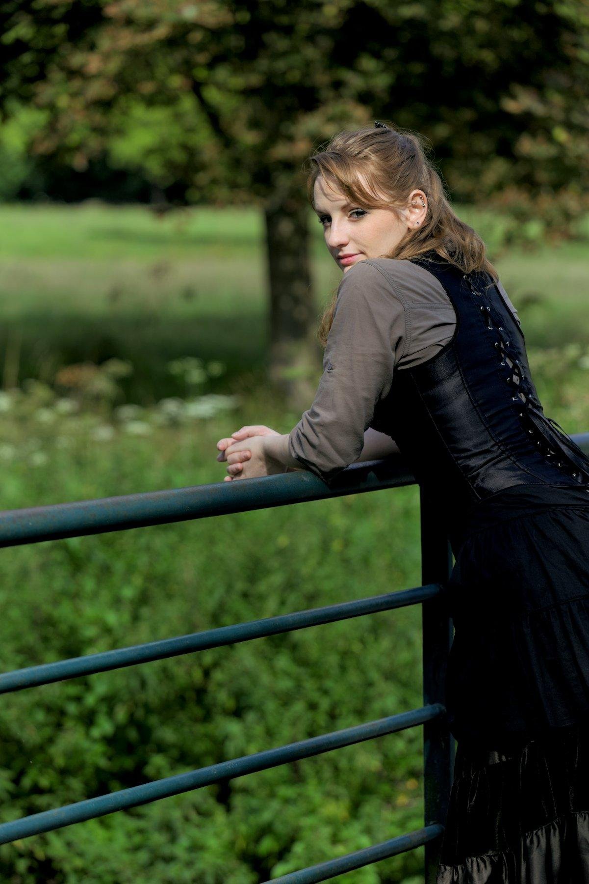 Photo portrait, montrant une jeune femme rousse au cheveux long élégamment coiffé, portant un chemisier marron et par dessus, un corset noir, une longue jupe marron foncé. Elle est accoudée à une rambarde en métal vert et regarde vers la caméra par dessus son épaule gauche