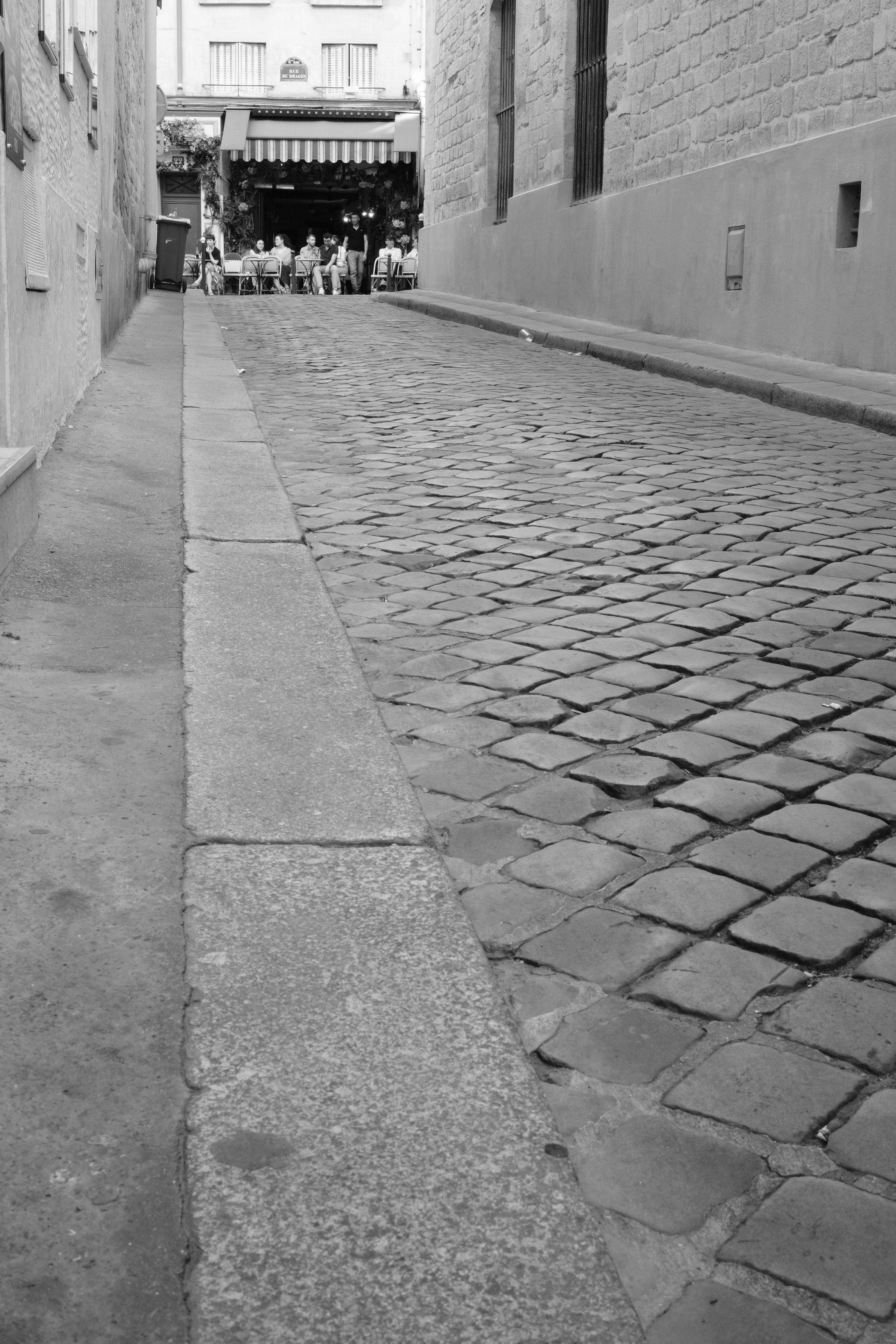 A photo taken very near from the floor, depicting a small paved street with buildings on both sides and going up to a pub with people sitting outside