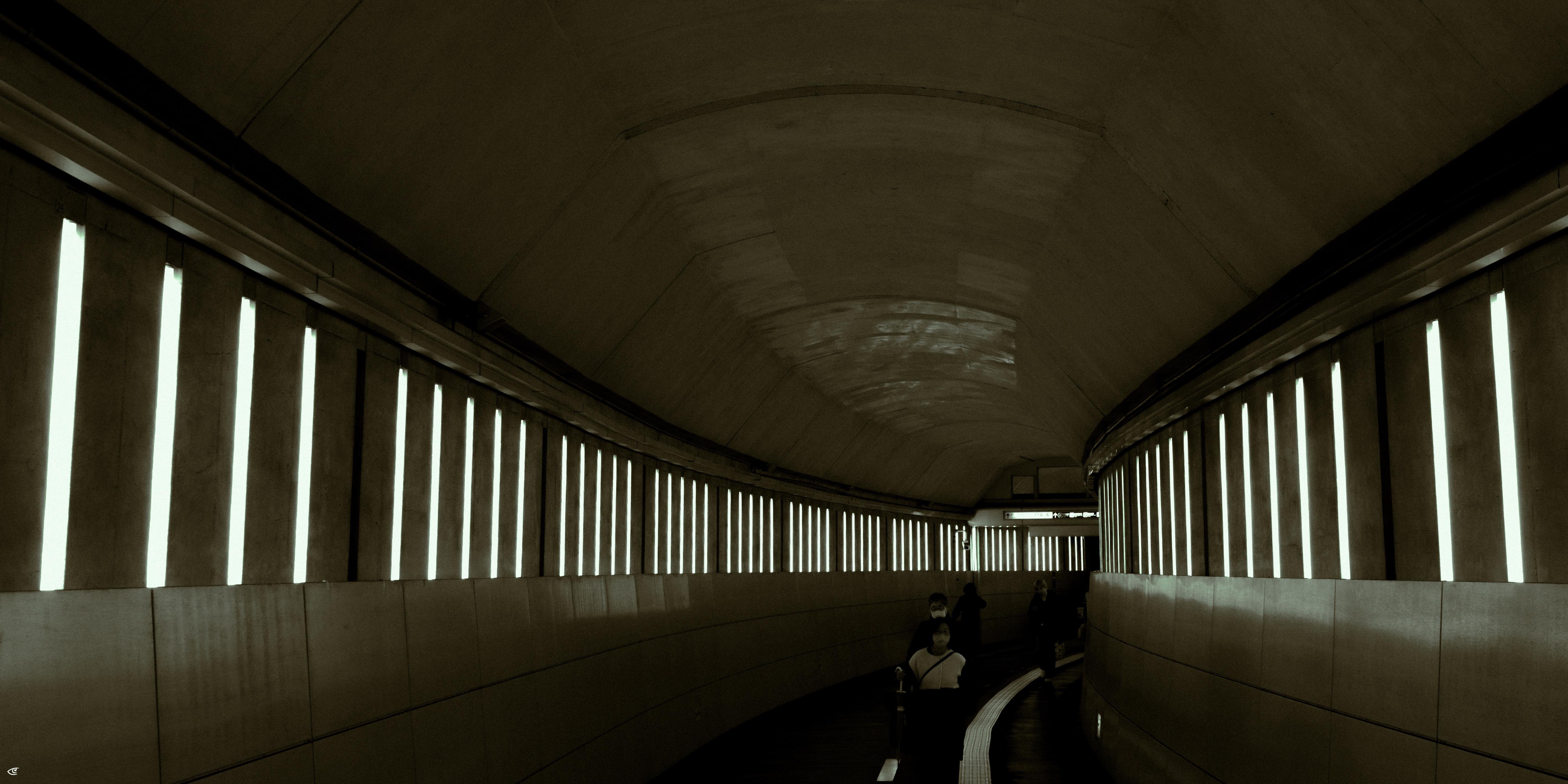 Curved underground tunnel with rows of vertical light panels along both walls, viewed from inside looking toward a bend; a few people are visible in the distance near the right side, with a narrow path and railing along the right wall.
