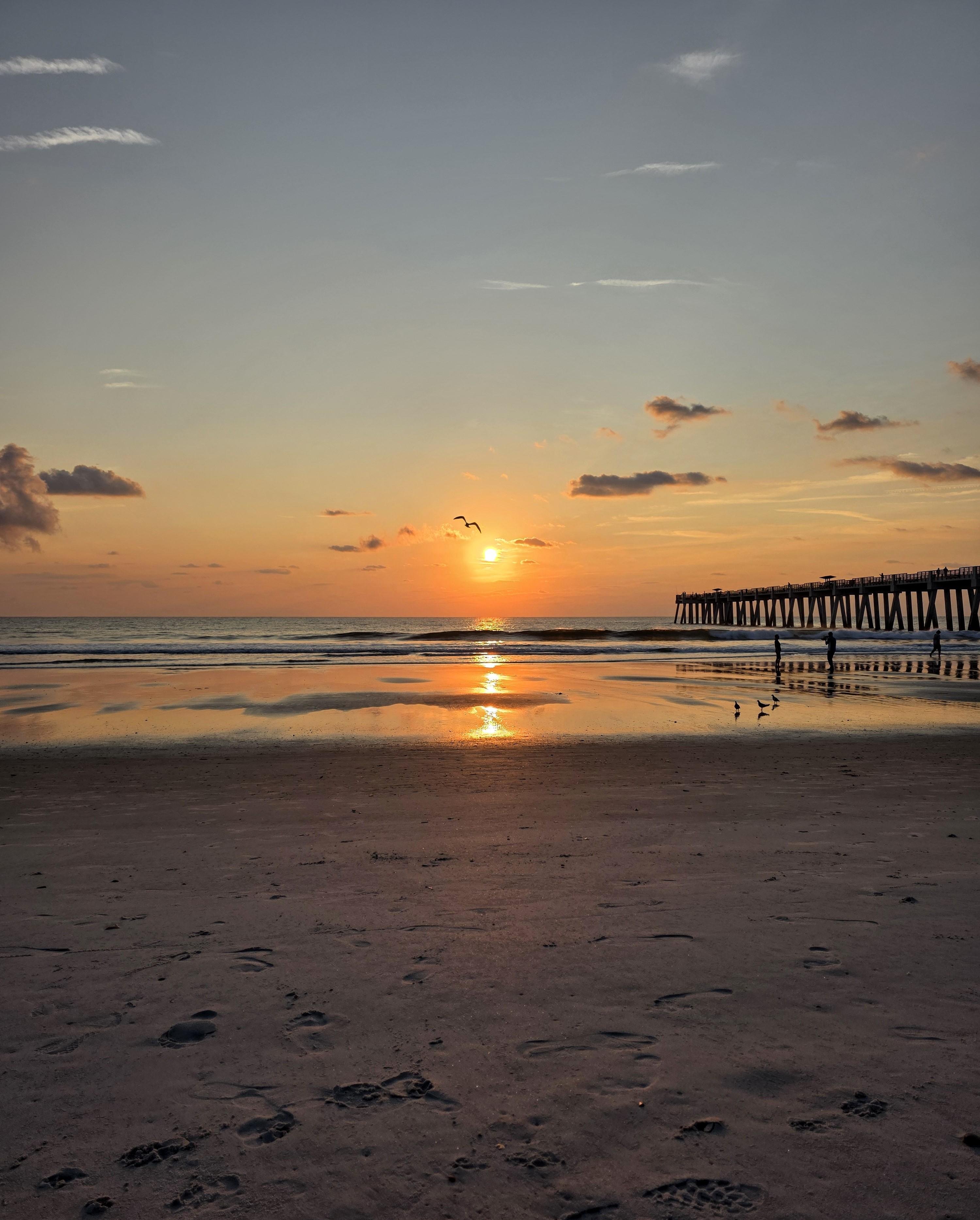 Sunrise over a beach with gentle waves reflecting the orange and pink hues of the sky. A long pier extends from the right, silhouetted against the horizon. Several birds fly near the sun, while a few are visible on the wet sand below. Shadows and footprints mark the foreground sand.