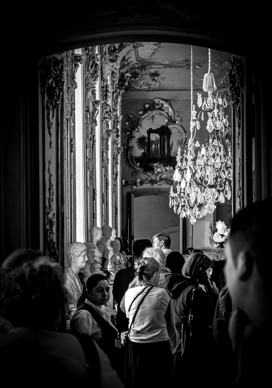 Crowded interior of an ornate hall with carved wall panels, a large crystal chandelier, and several classical busts on pedestals along the left wall; people stand and walk beneath a decorative mirror and archway.