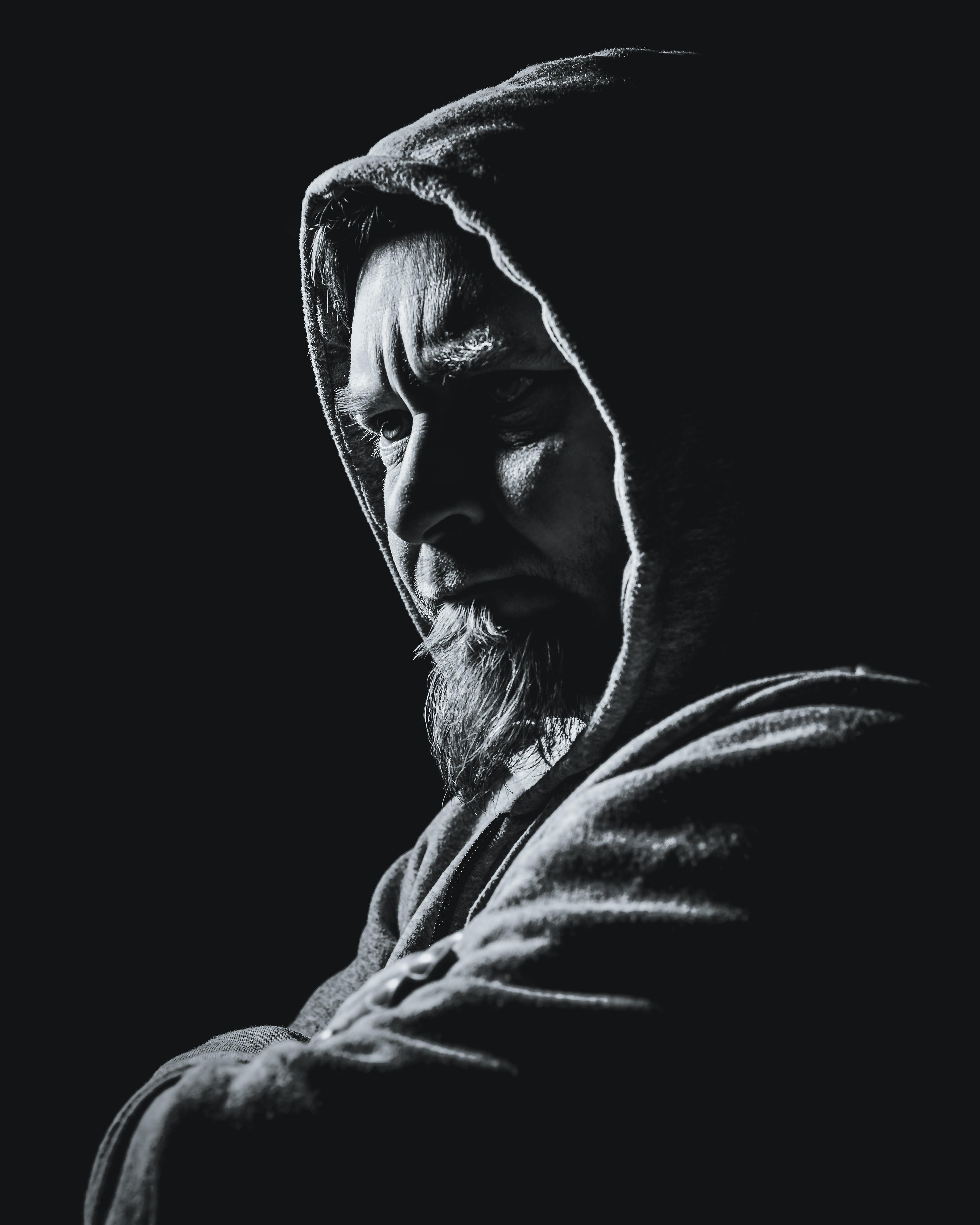 The image is a black-and-white portrait of a person wearing a hooded sweatshirt. The lighting creates dramatic shadows on their face, emphasizing their features. The person's expression appears contemplative or introspective.