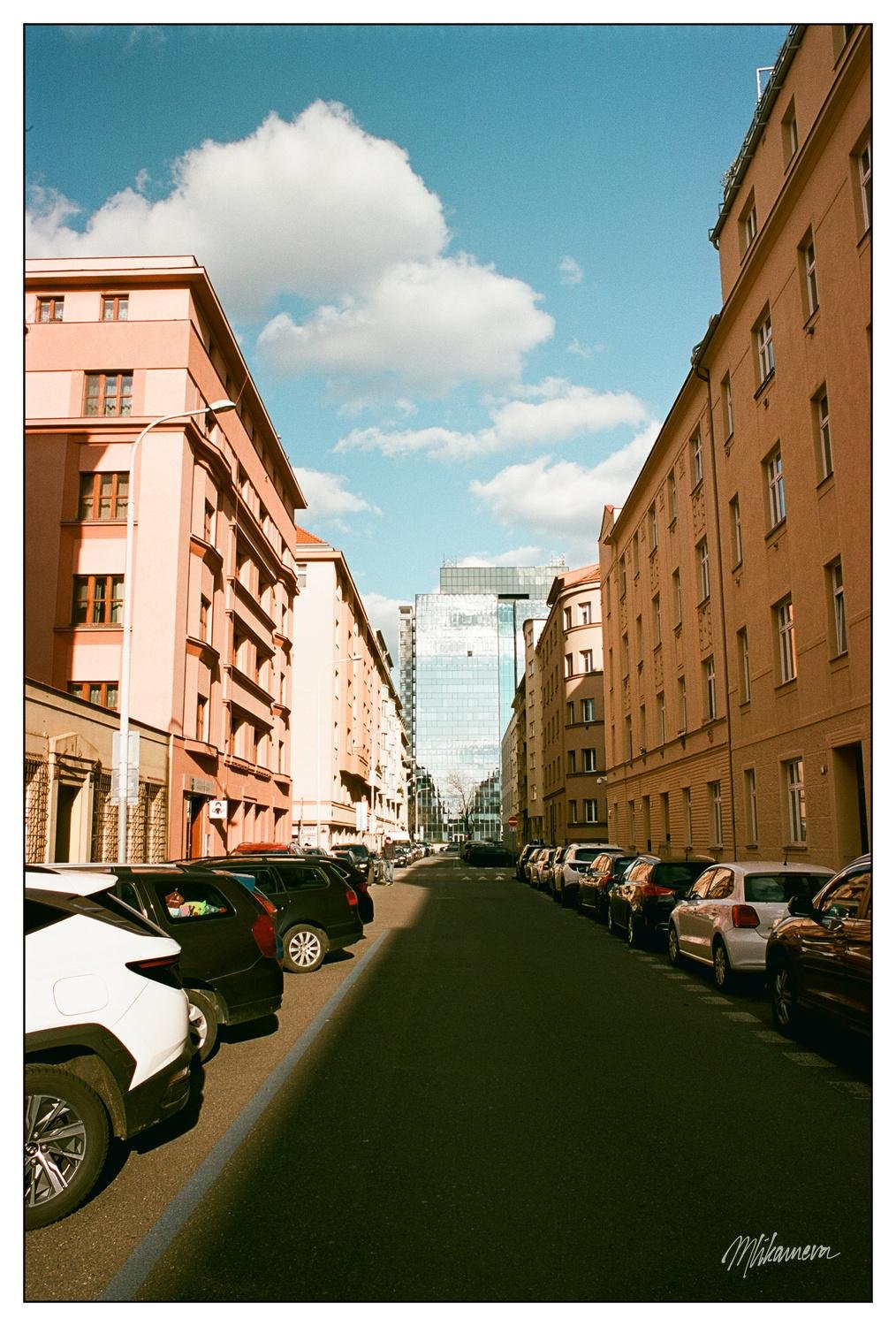 Street scene with parked cars lining both sides, bordered by tall buildings with red and beige facades. In the background, a modern glass skyscraper reflects sunlight. Blue sky with scattered white clouds.