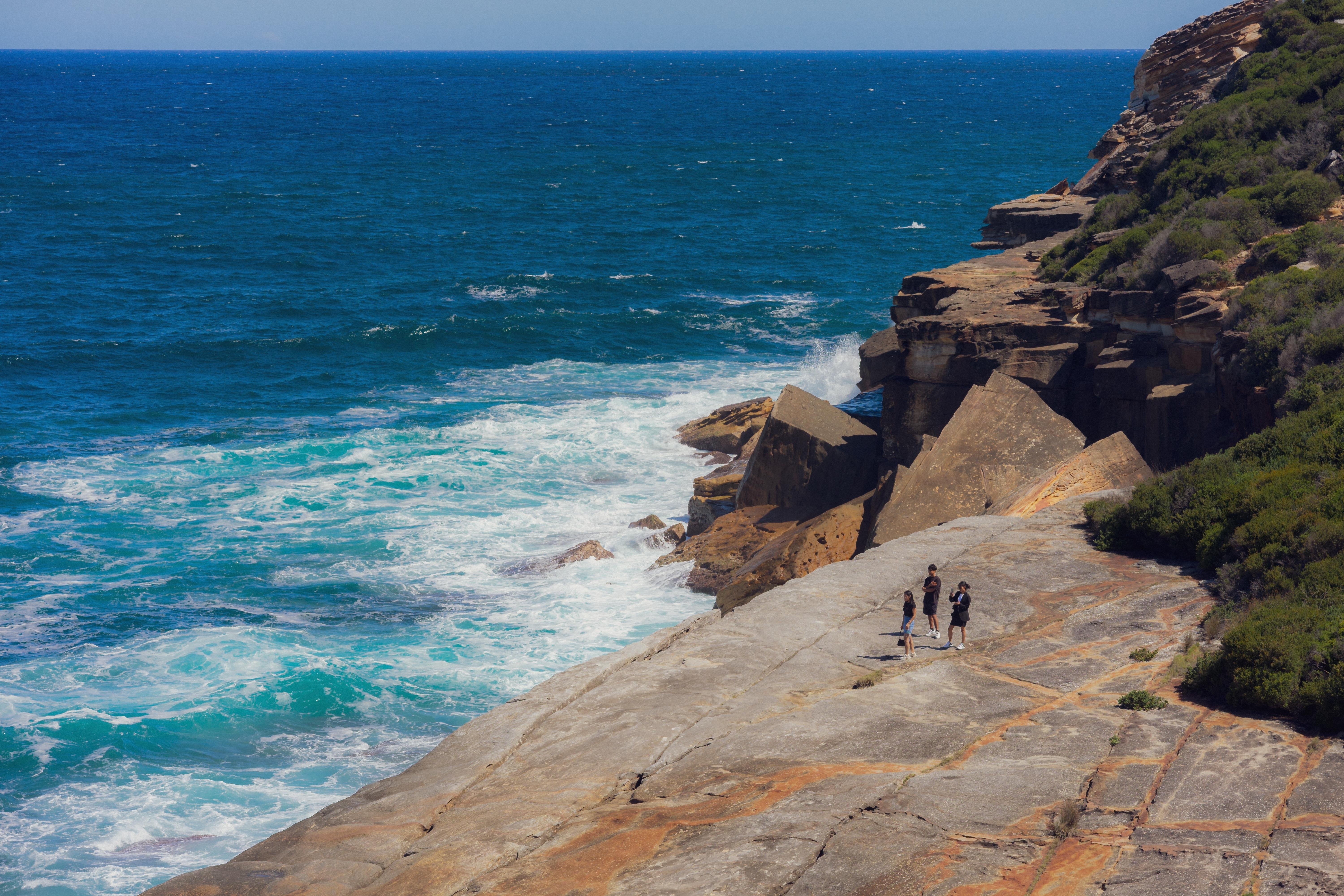 The image shows a coastal scene with a rocky cliff and an ocean view. There are three people walking on a flat rock surface close to the water. The waves are crashing against the rocks, and there’s vegetation on the right side of the image. The sky is clear, indicating a sunny day.