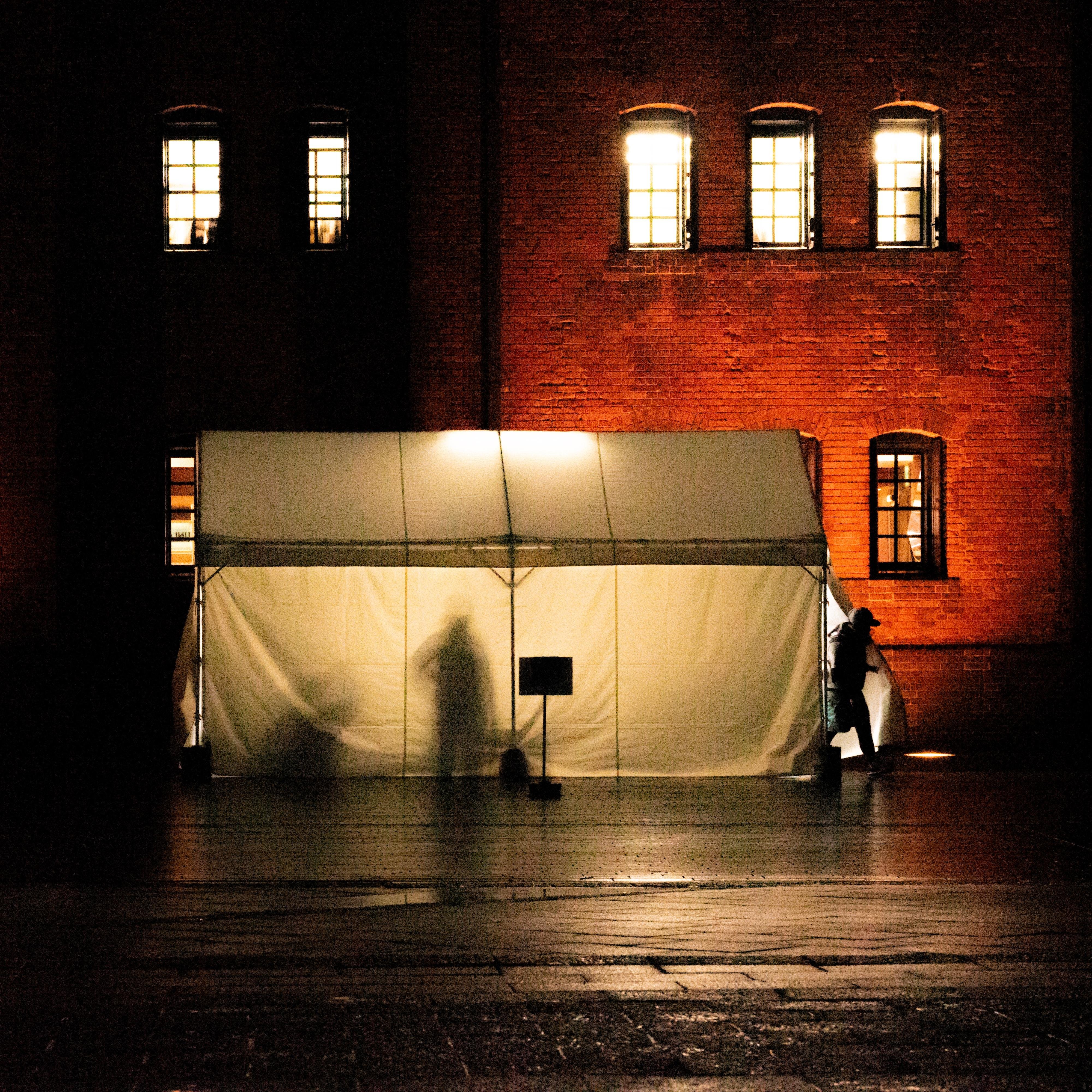 A white tent is illuminated from inside, casting shadows of people on its fabric. The tent is set against a red brick building with lit windows. A person is visible near the tent's entrance. The ground is dark and appears wet, reflecting light from the surroundings.