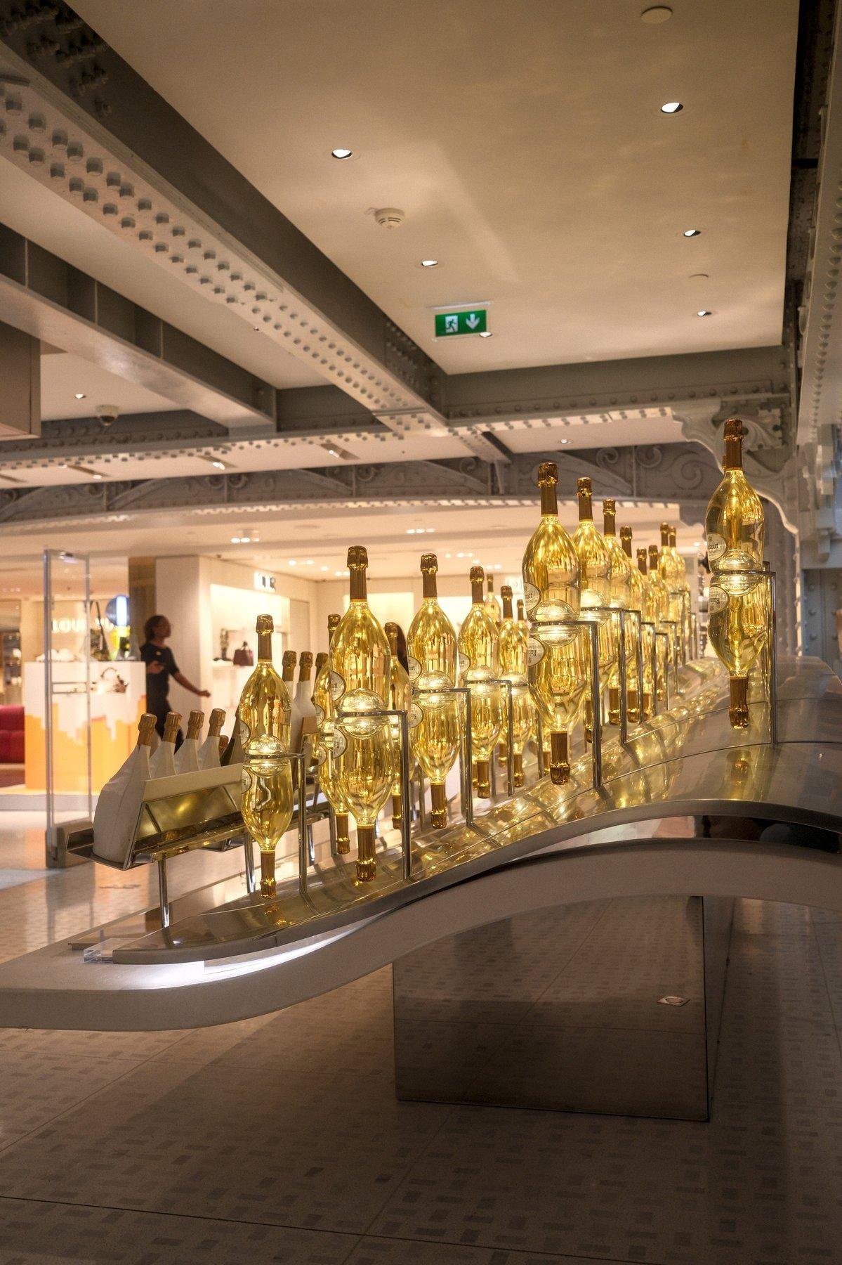 Photo verticale. Sur un présentoir formant une petite vague, des bouteilles d’alcool jaune sont alignées, avec à chaque fois, une bouteille à l’endroit posée sur une bouteille à l’envers