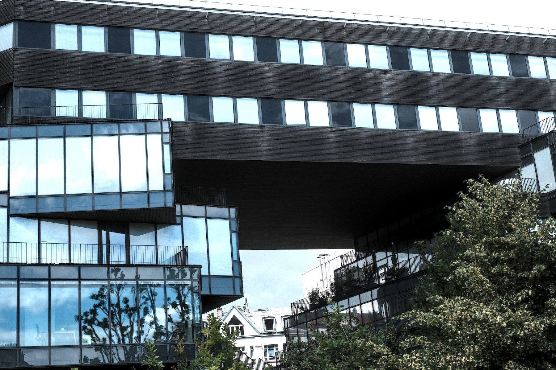 Photo horizontale. Un bâtiment en verre et acier dont les étages sont comme posés les un sur les autres sans être alignés. Dessus, comme posé négligemment, un bâtiment de 2 étage à la façade en bois noir et assez large, fait la passerelle vers un autre bâtiment en verre et acier
