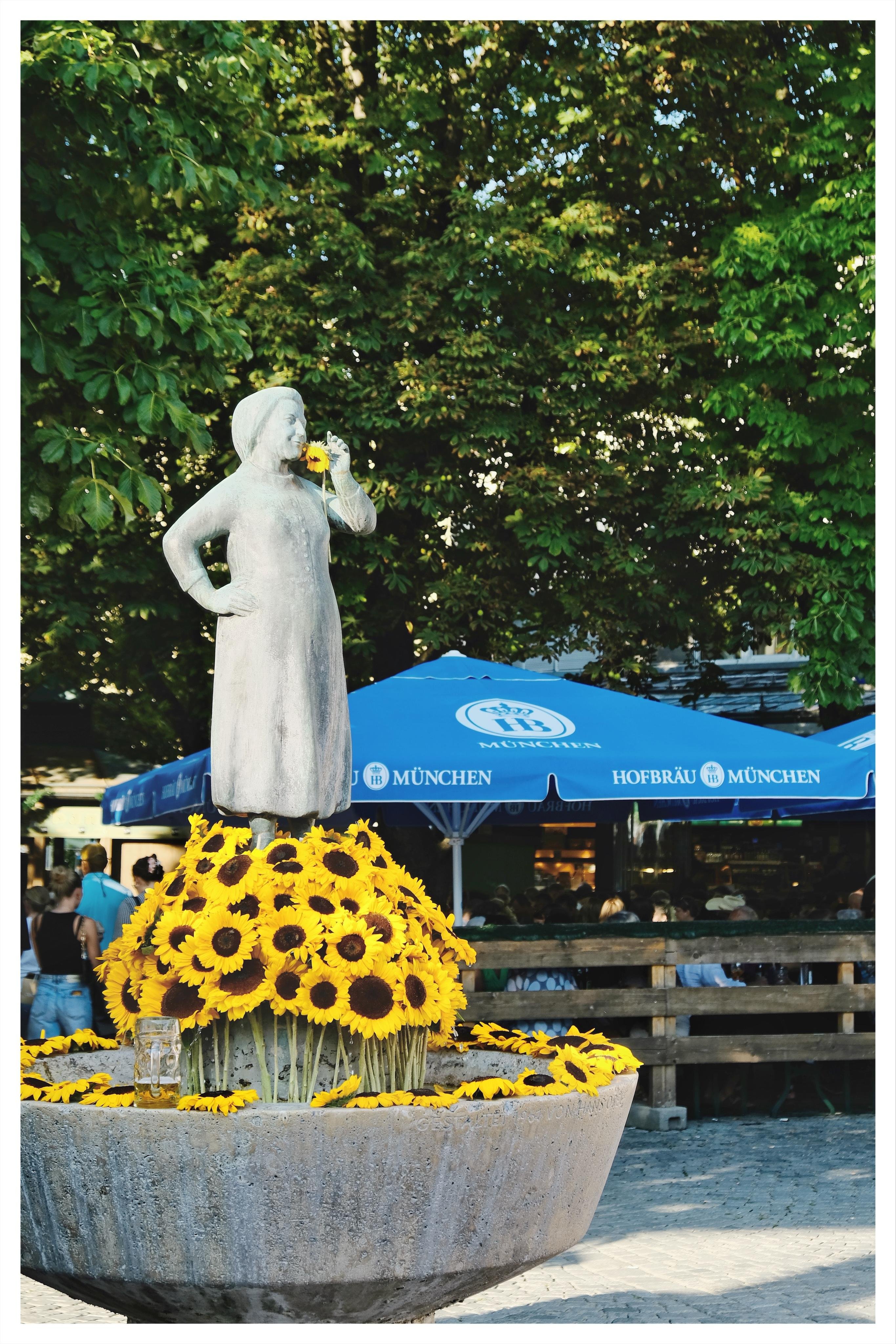 Gray statue of a woman holding one hand near her mouth, standing on a large stone planter filled with bright yellow sunflowers; blue umbrellas in the background read “Hofbräu München,” with people seated beneath and leafy trees overhead.