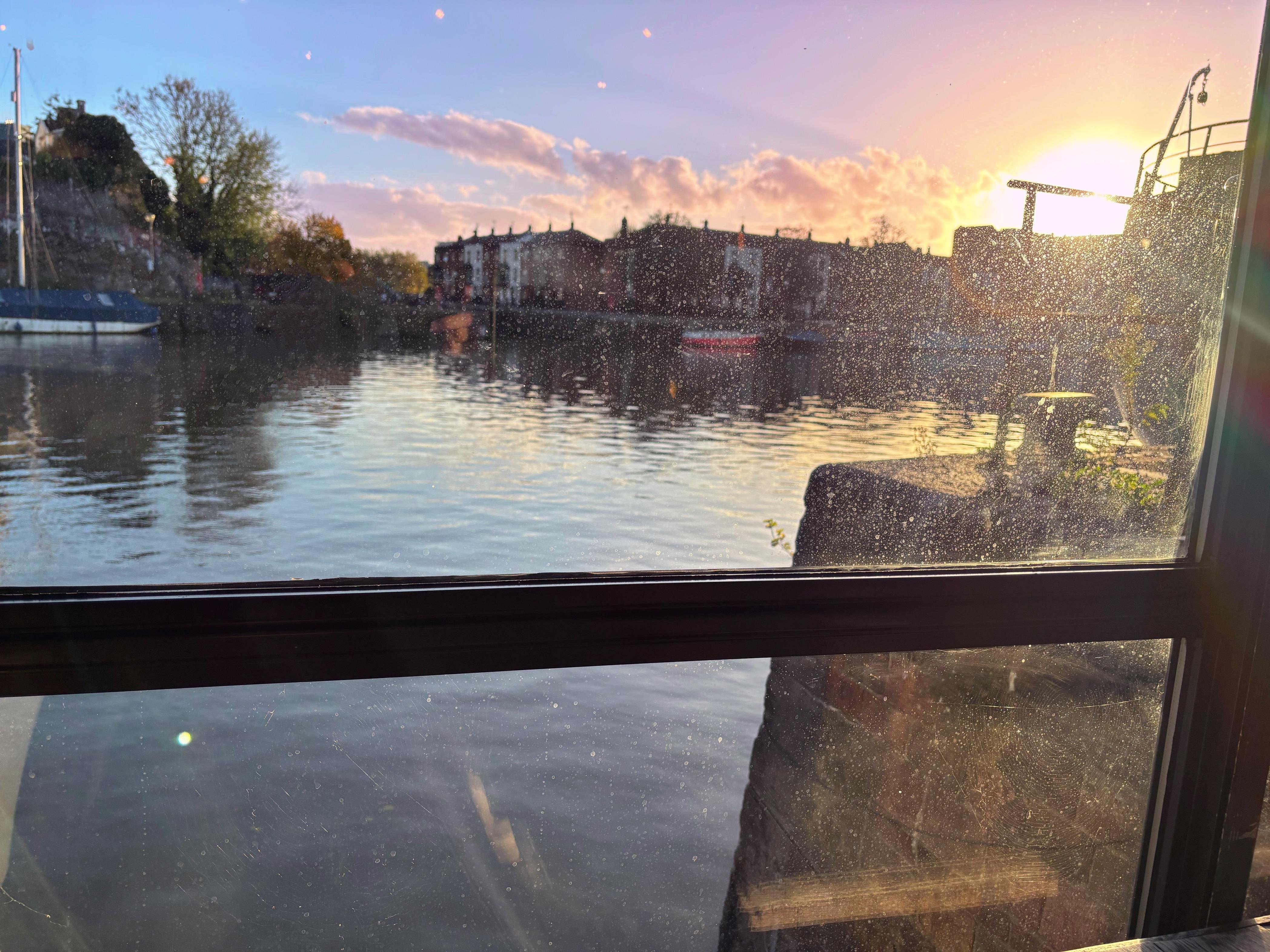 The image shows a view through a window overlooking a body of water, likely a river or canal. There is a boat visible on the left side and buildings in the distance. The sun is setting, casting a warm glow across the scene, and there are reflections on the water. The window appears to have some dust or water spots on it.