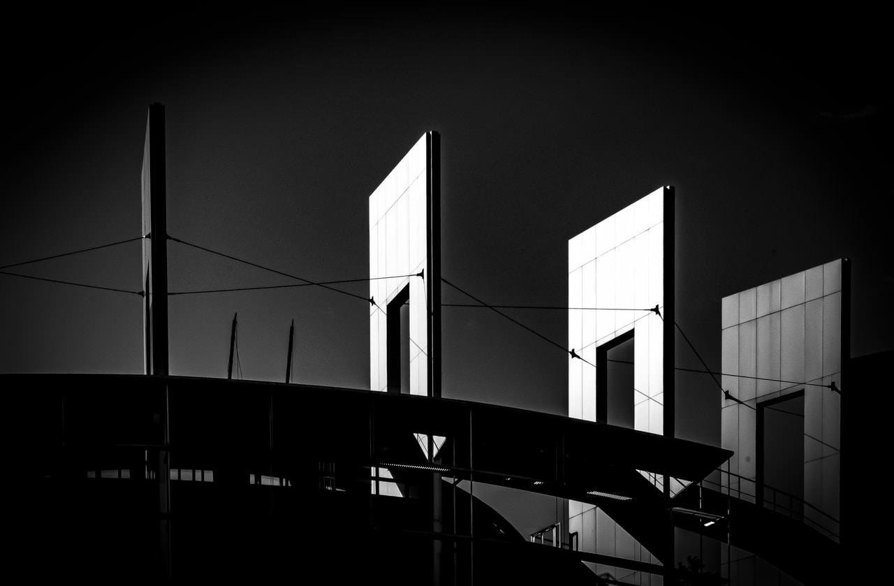 Three illuminated rectangular panels stand vertically against a dark backdrop, supported by cables. The foreground contains a curved structure, partially silhouetted, with visible railings and beams. The overall composition presents strong contrasts in black and white.