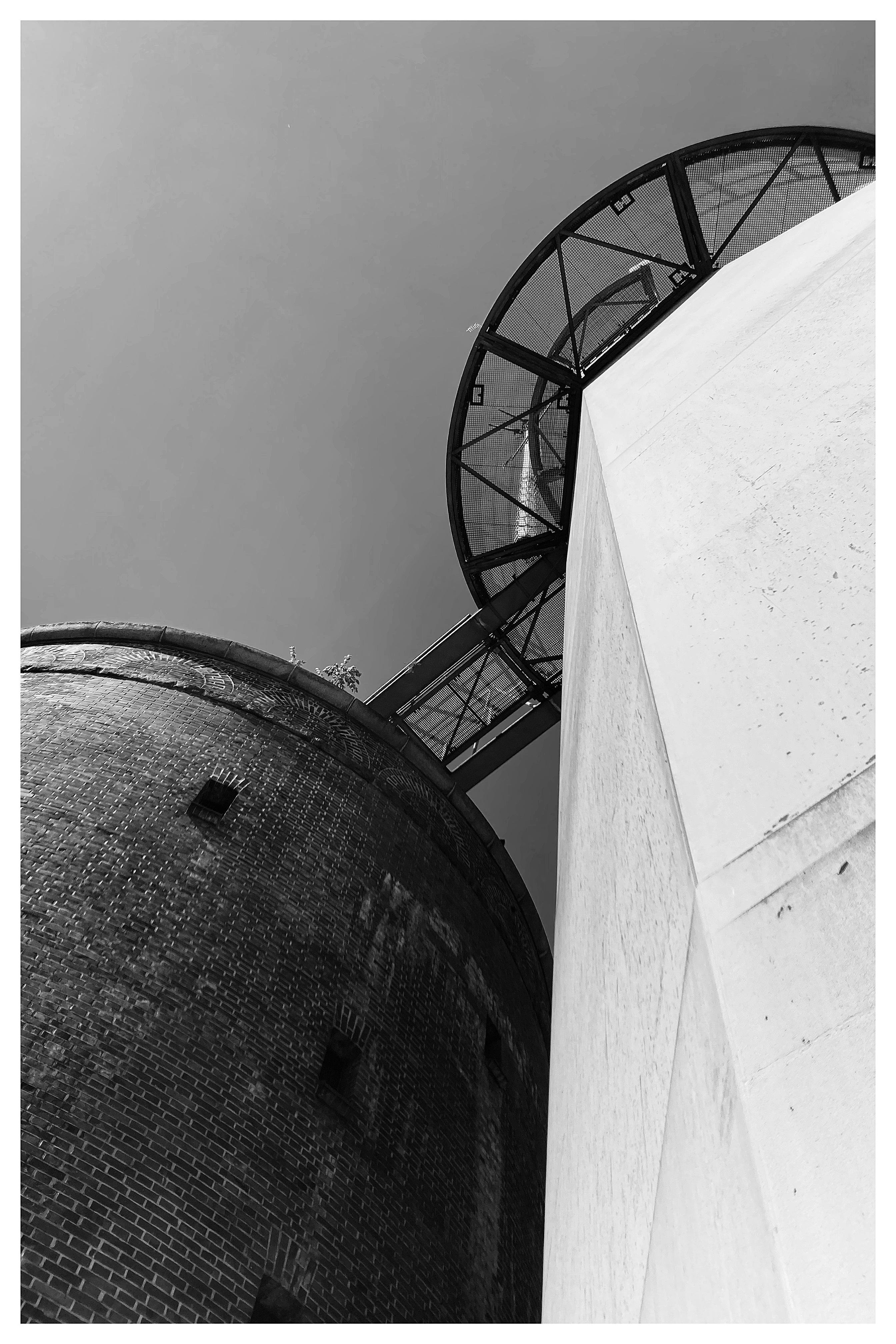 Low-angle black and white view of two adjacent cylindrical structures, one with a brick surface and the other with a smooth, light-colored finish. The upper part of the image shows a circular metal platform with fencing and structural beams. The sky is clear and light grey. Shadows and textures are visible on the surfaces.