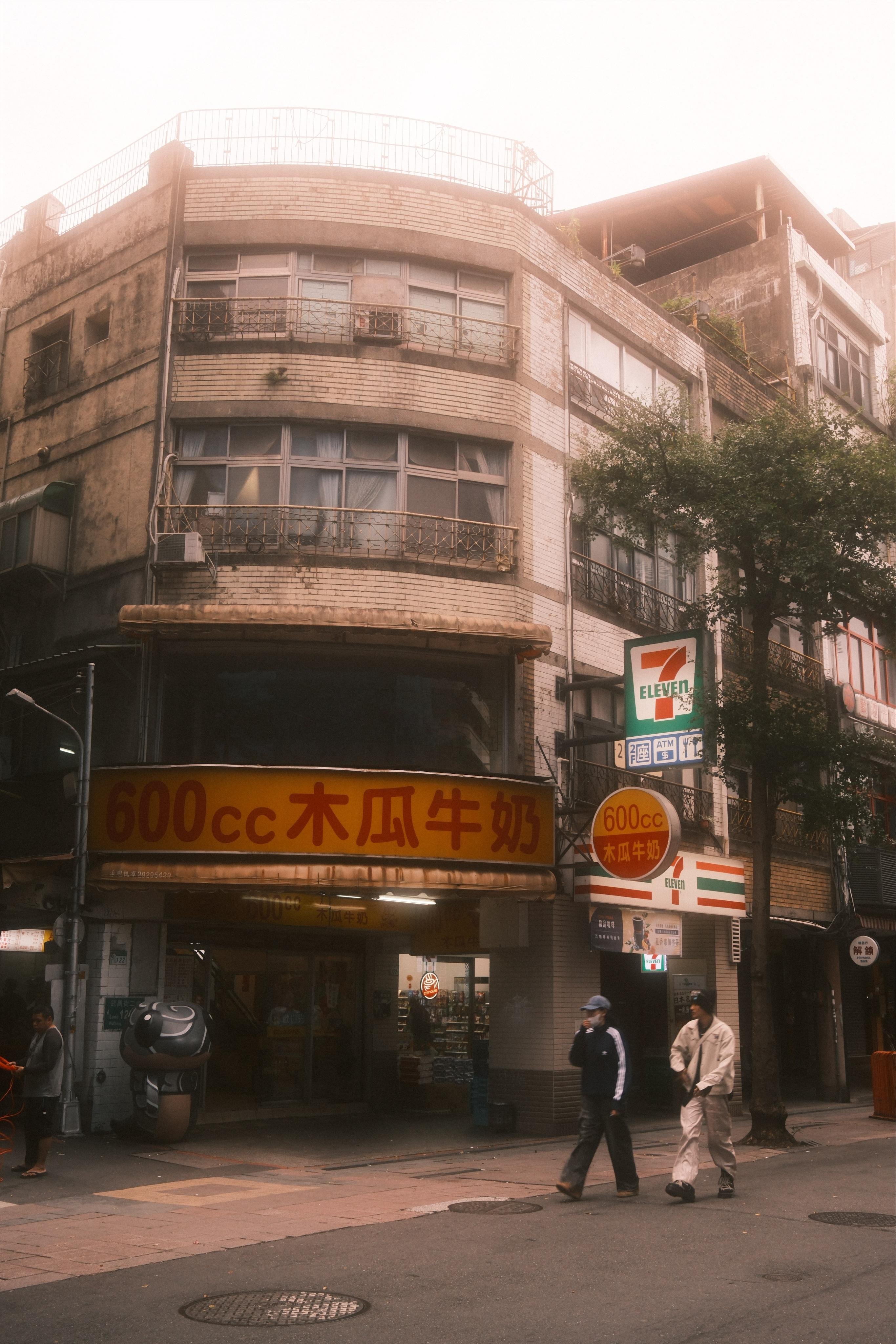 Street corner with a curved, multi-story brick building featuring bay windows and metal balcony railings; a 7‑Eleven sign and storefront at ground level beneath a large yellow sign with Chinese characters and “600cc,” with a few pedestrians walking along the sidewalk and trees partly covering the facade.