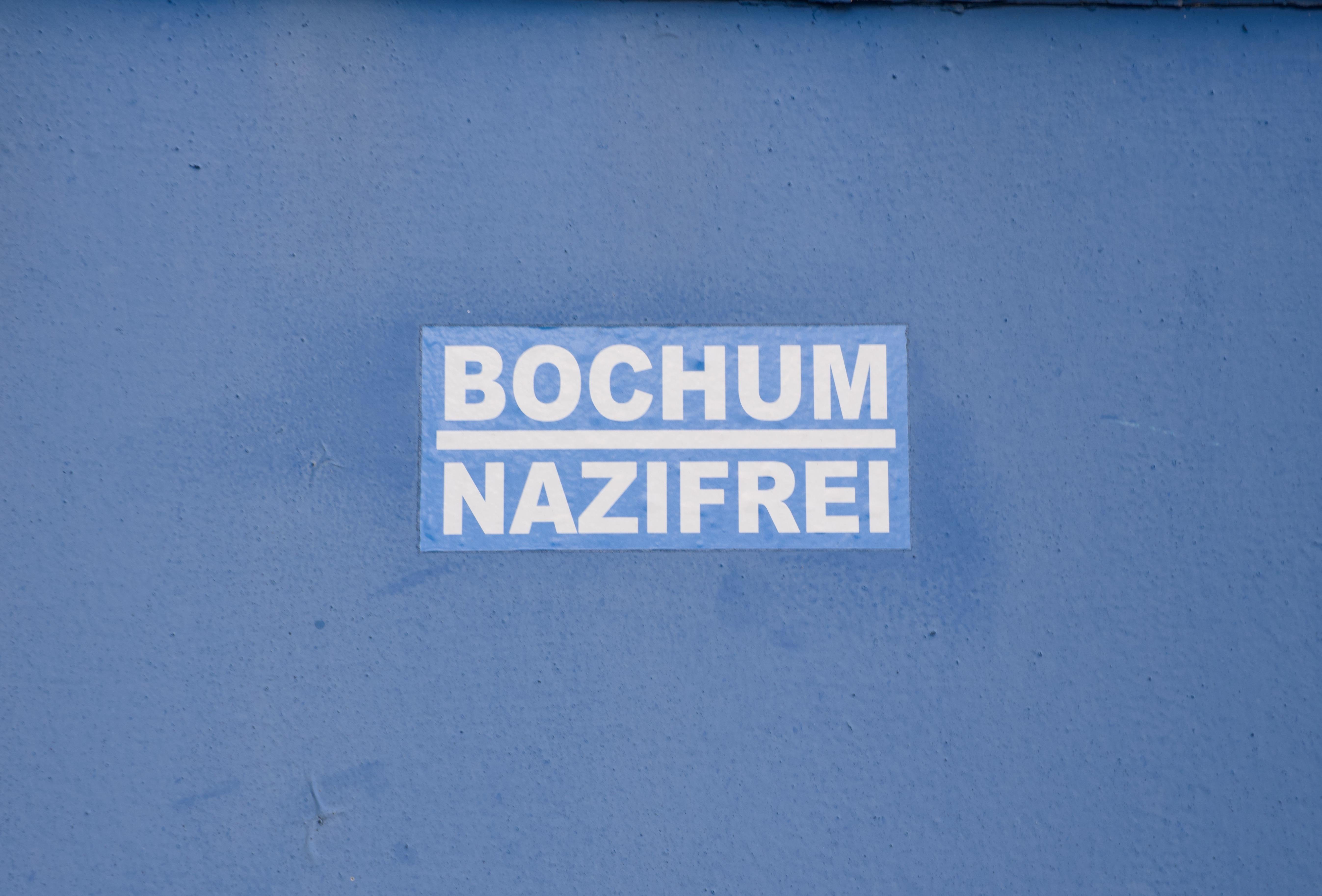 A blue wall features a white sticker with bold text in two lines reading "BOCHUM NAZIFREI," with a white underline separating the words. The wall's surface is smooth, with slight imperfections visible.