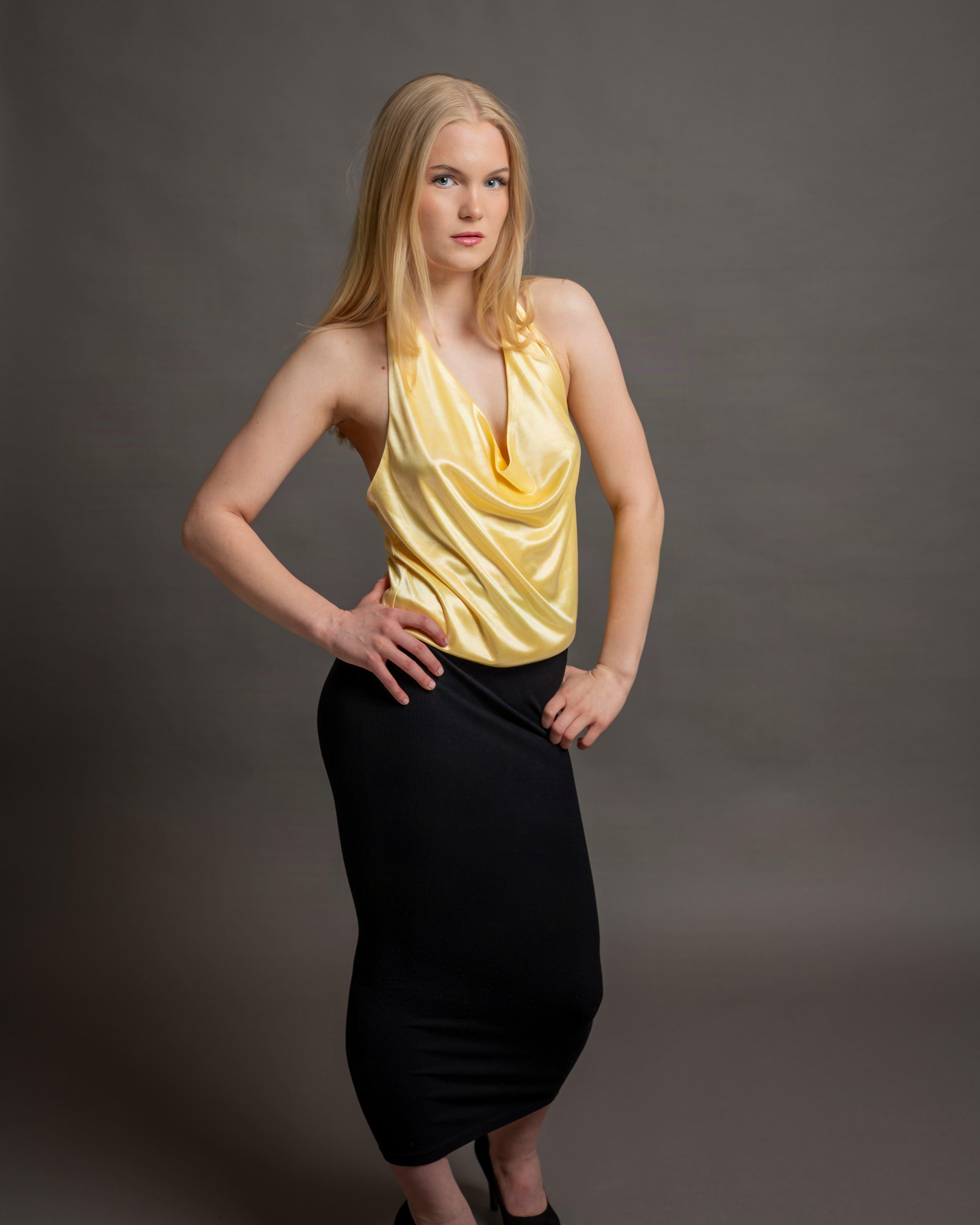 Woman with long blonde hair wearing a yellow satin halter top and a black high-waisted, form-fitting skirt. Her arms are akimbo, with hands resting on her hips. She stands against a plain gray backdrop, gazing directly at the camera.