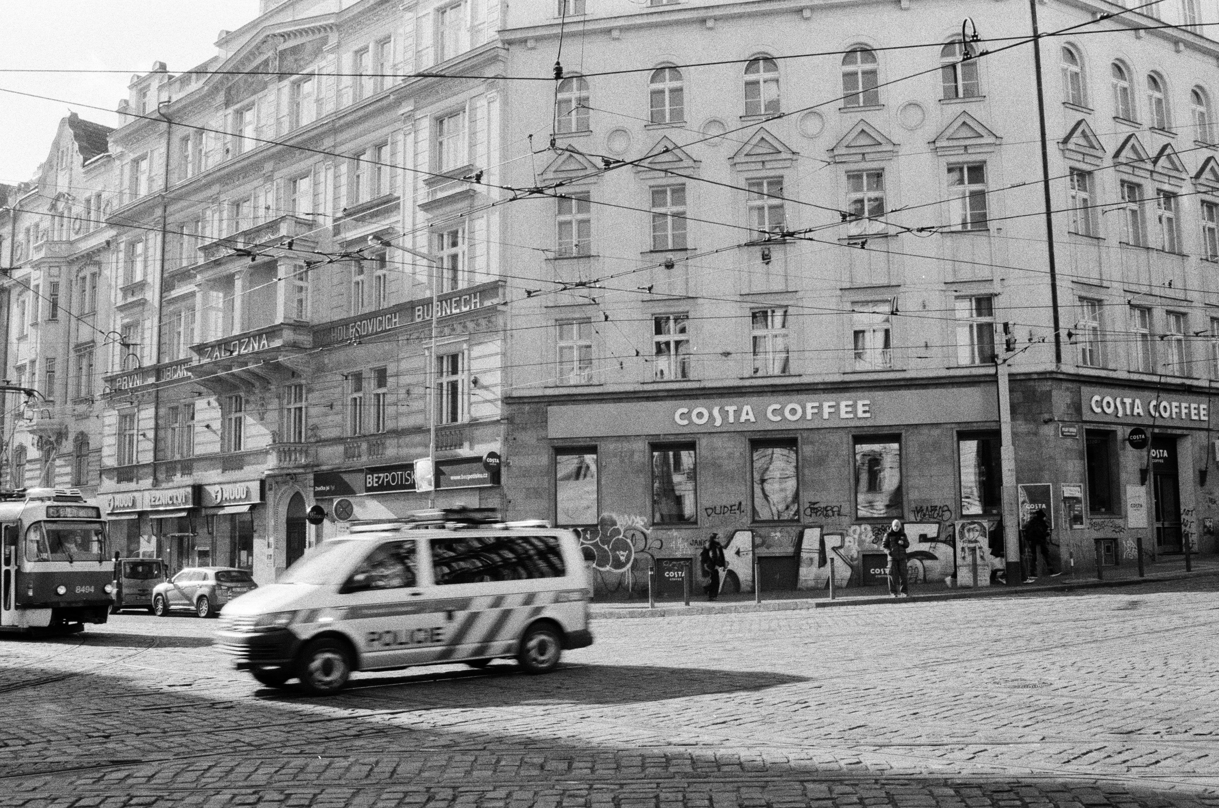 The image is a black and white street scene featuring a corner building with a Costa Coffee shop. There's a police vehicle in motion and a tram in the background. The building is adorned with various signs and has graffiti on the lower part. It's an urban setting with cobblestone streets and overhead wires, likely for power lines or public transportation.