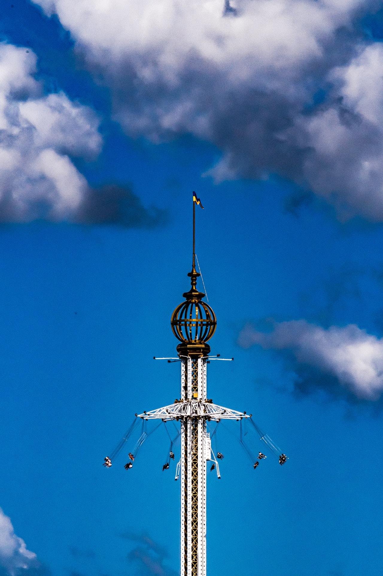 Tall amusement park ride with suspended swing chairs rotating around a central column, topped with a decorative metal globe and small flag. Bright blue sky with scattered white clouds in the background.