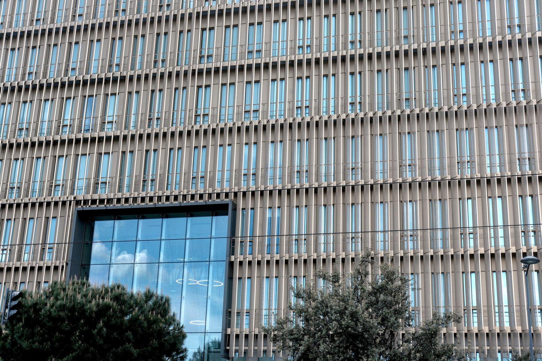 Photo horizontale. Un grand bâtiment moderne en verre avec des barreaux en bois clair sur toute la façade, Une grosse découpe dans le bois est présente au niveau de l’entrée du bâtiment