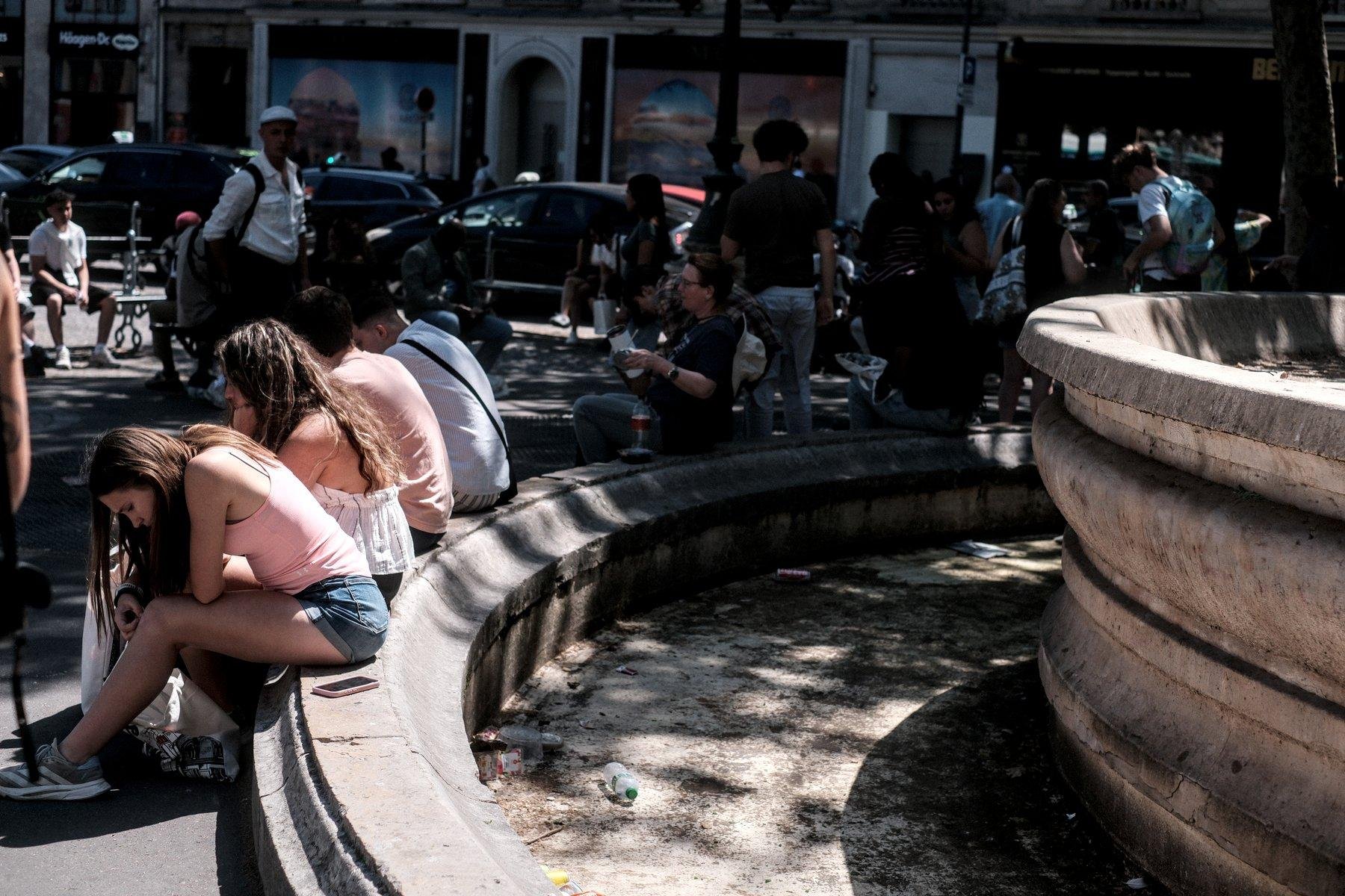 Photo horizontale. Vue sur un bout d’une fontaine circulaire, mais éteinte et vide. Des gens sont assis sur le bord.