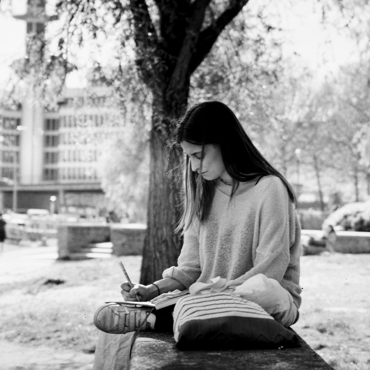 The image shows a black and white photograph of a person sitting outside, drawing in a notebook. They are sitting on a bench with a tree and some buildings in the background. 