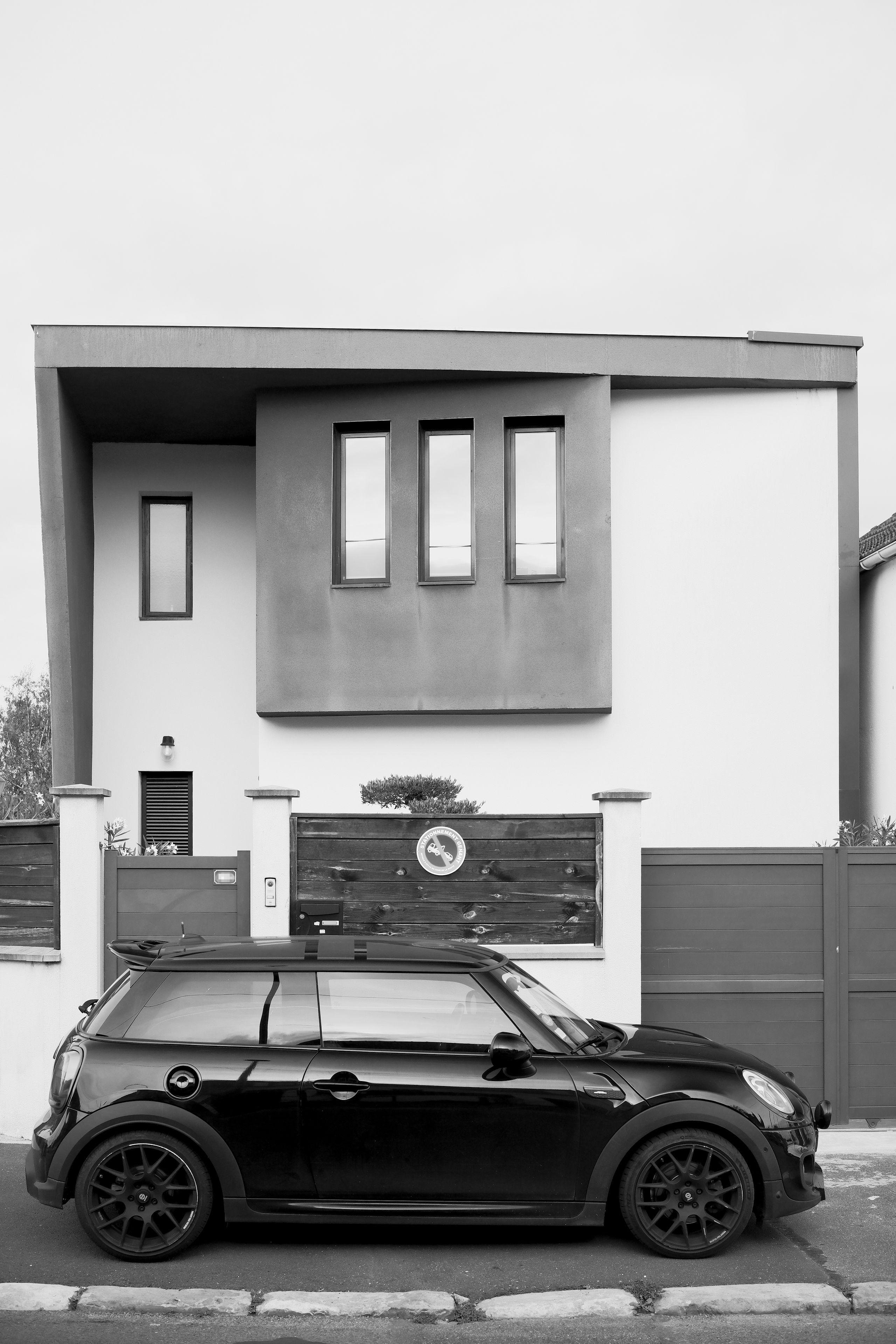 Photo verticale en noir et blanc. Une petite voiture noire garée devant une maison moderne où on peut voir un panneau interdiction de stationner accrocher à la palissade