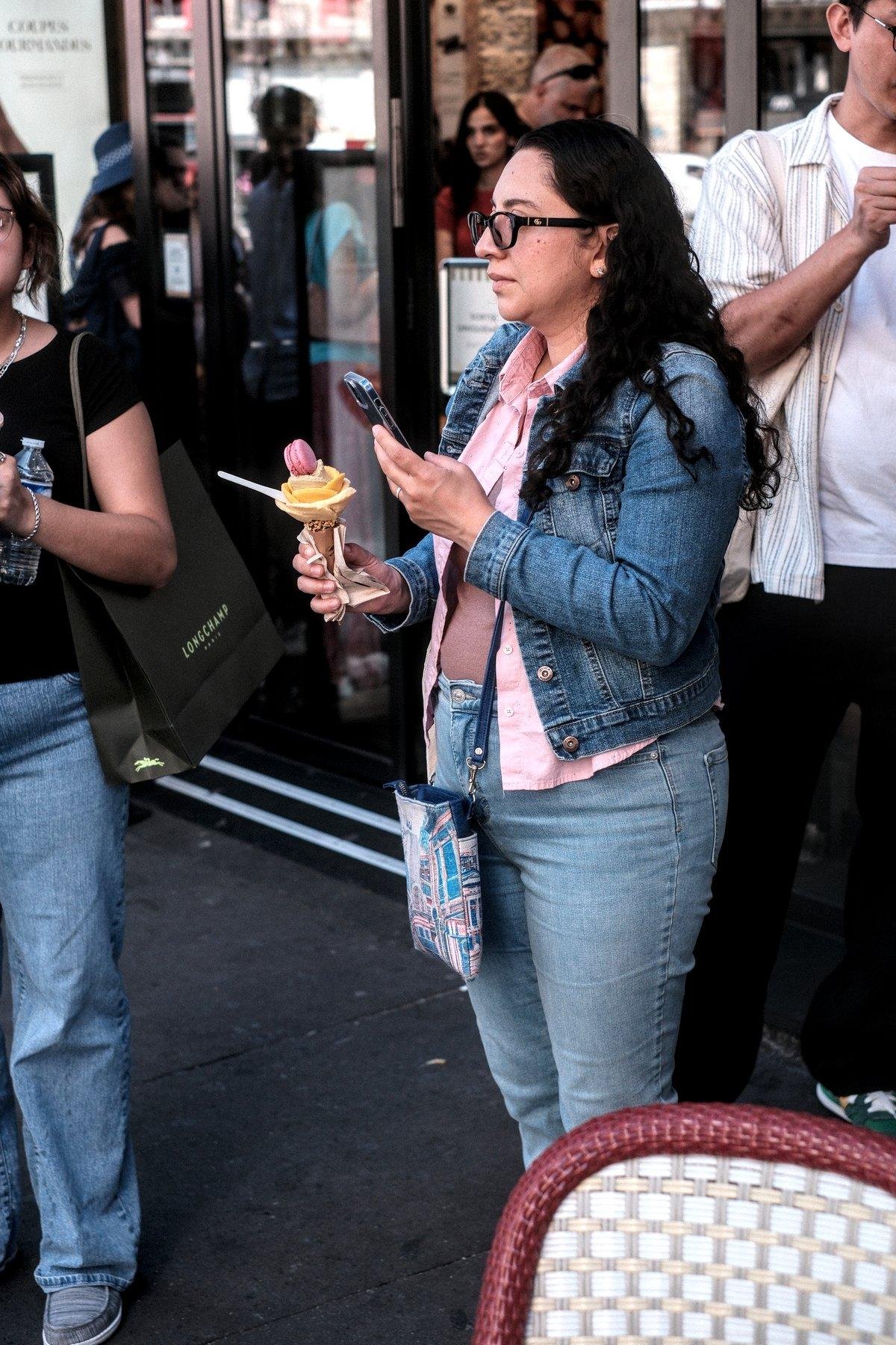 Photo verticale. Une femme brune cheveux longs lâchés, veste en denim sur chemise rose ouverte sur débardeur marron, jean bleu, prenant en photo son cône de glace en forme de fleur avec un macaron au sommet