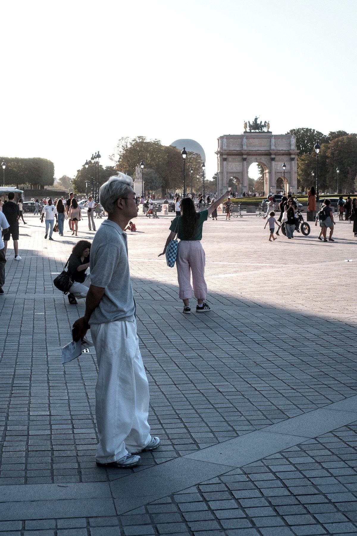 Vertical photo. On the Louvre’s place, a young man profile facing the right side of the pic, with silvery hairs is waiting for something or someone, arms in the back holding a flyer. Behind, a woman is doing the “pinch the Louvre pyramid” pose and her friend is crouching to take the photo. Half of the pic is in the shadows and the other half under the sun