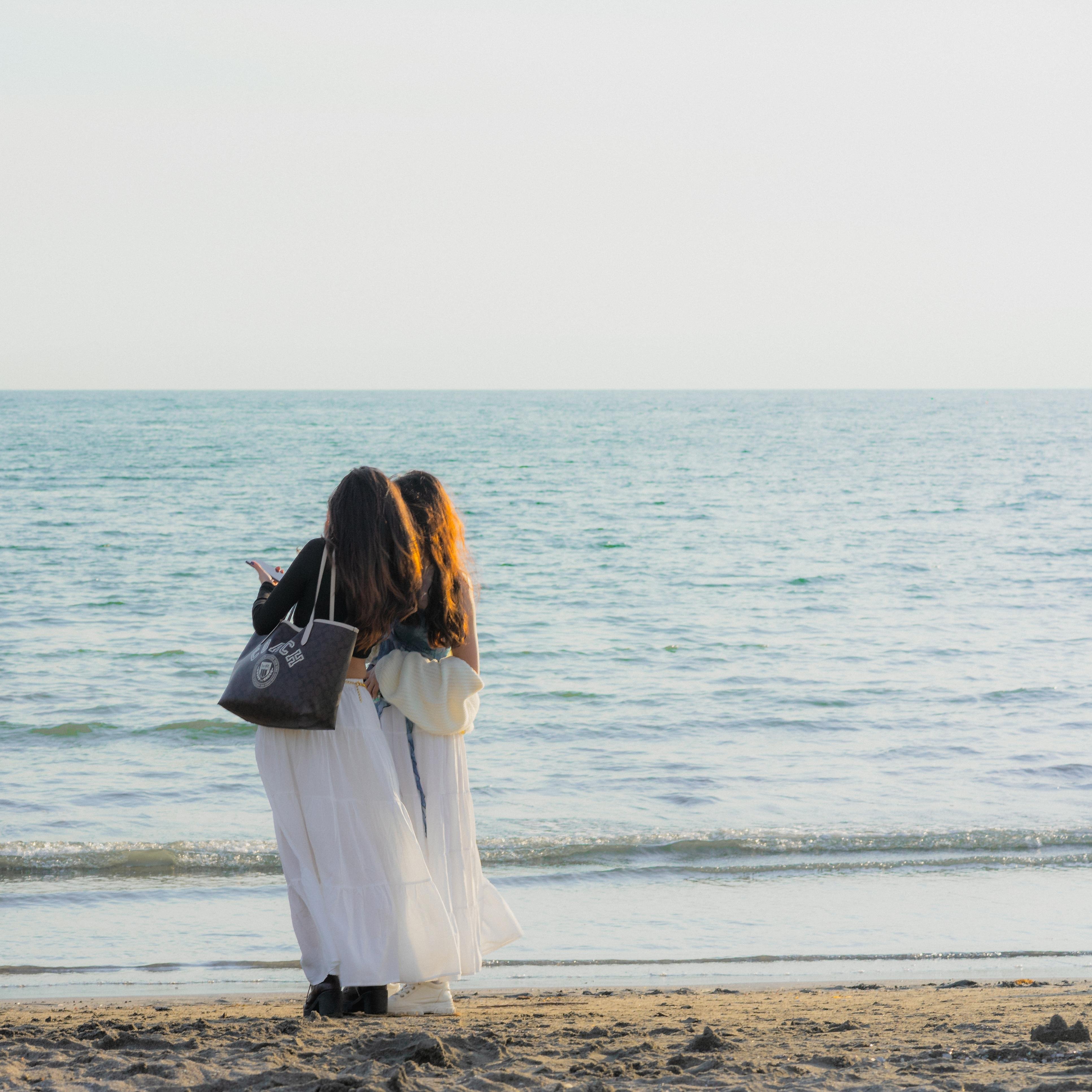 The image shows two people standing on a beach facing the ocean. They appear to be close to each other, possibly embracing or posing. One of them has a tote bag. The setting is serene, with calm waves and a clear sky.