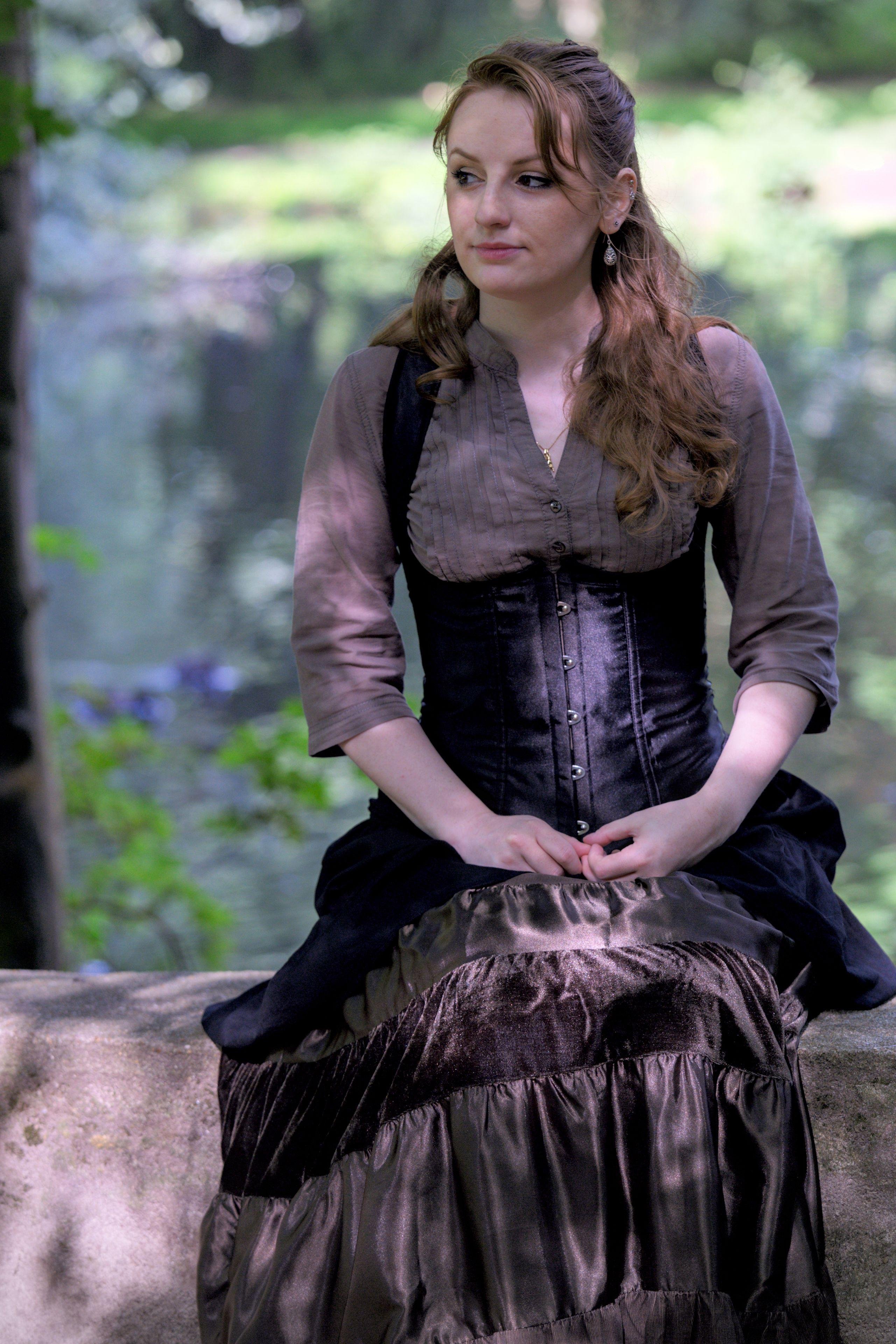 Photo portrait, montrant une jeune femme rousse au cheveux long élégamment coiffé, portant un chemisier marron et par dessus, un corset noir, une longue jupe marron foncé. Elle est assise sur un muret et ses mains se tortillent les doigts comme par nervosité mais elle a un petit sourire aux lèvres