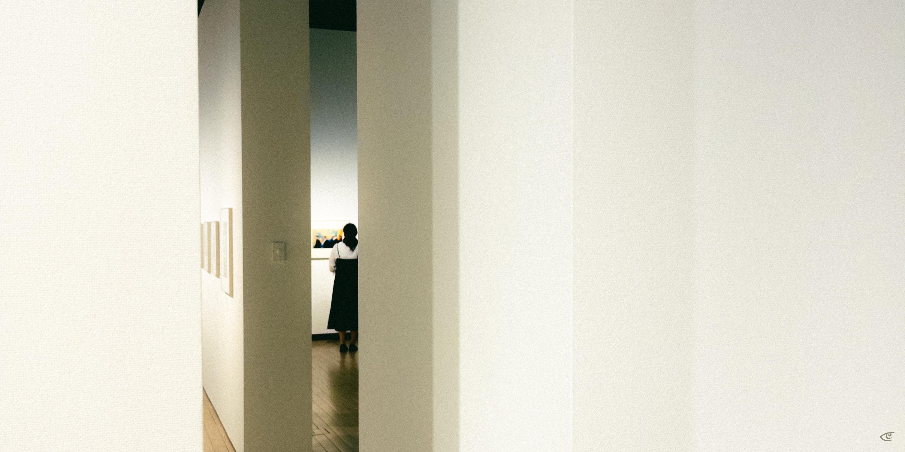View down a white-walled gallery corridor framed by tall rectangular pillars; a person stands in the distance near a brightly lit display, with small framed artworks on the left wall and a wood floor below.