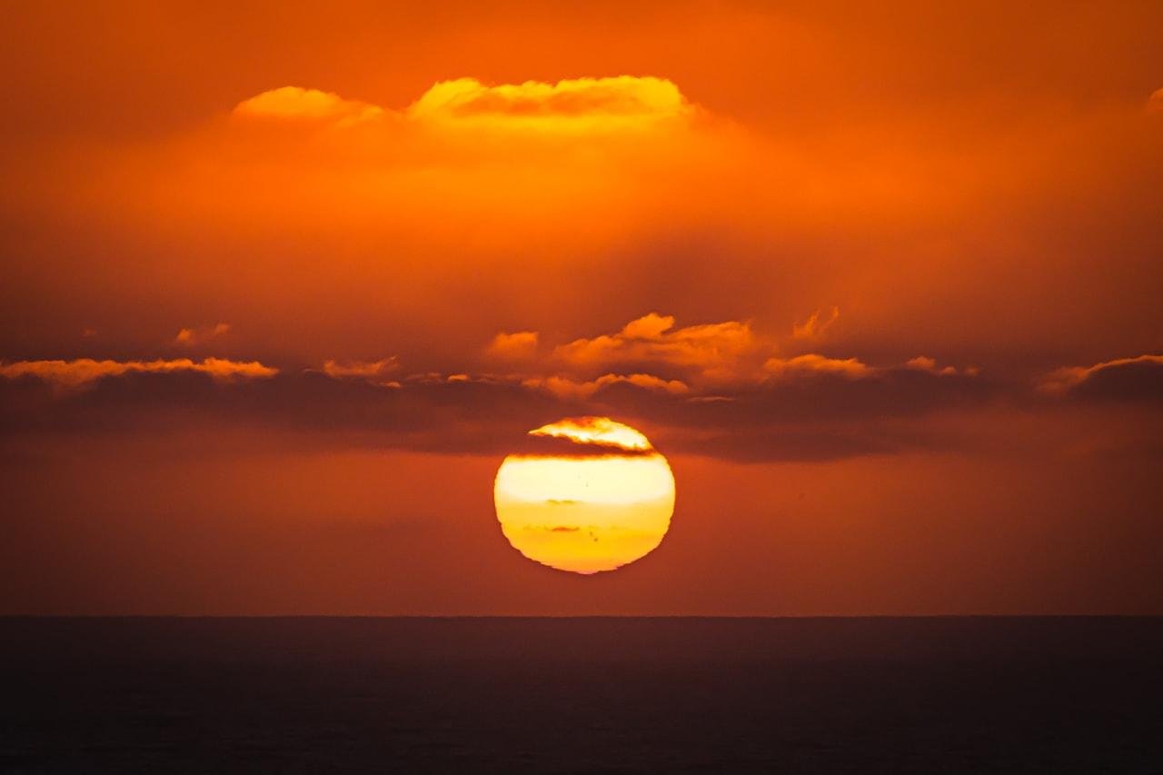 The image shows a sunset over the ocean, with the sun partially obscured by clouds. The sky is a rich orange color, and the reflection on the water creates a serene and warm atmosphere.