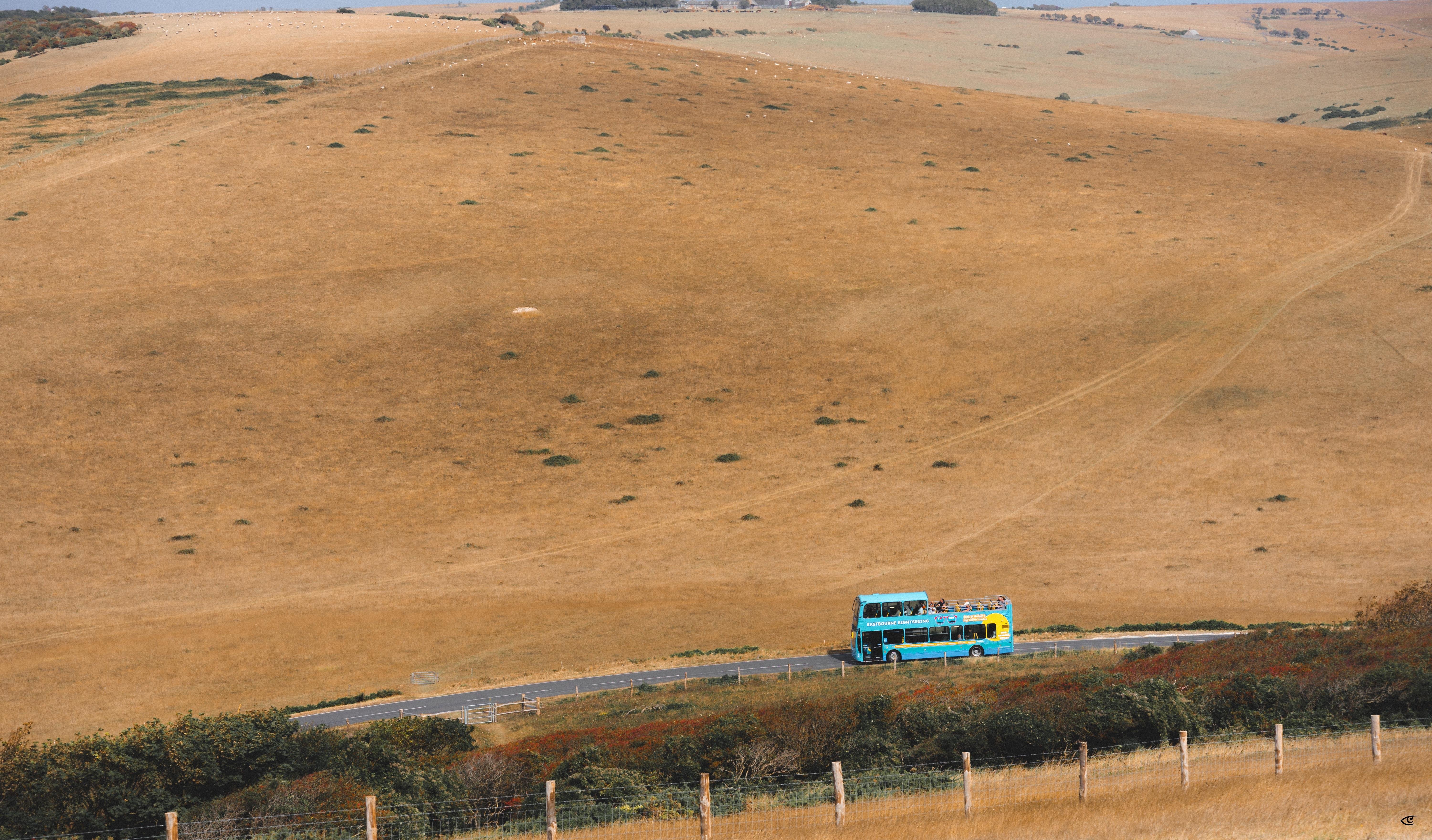 Turquoise double-decker bus traveling on a road at the bottom of the frame, with rolling tan hills and sparse vegetation stretching across the background.