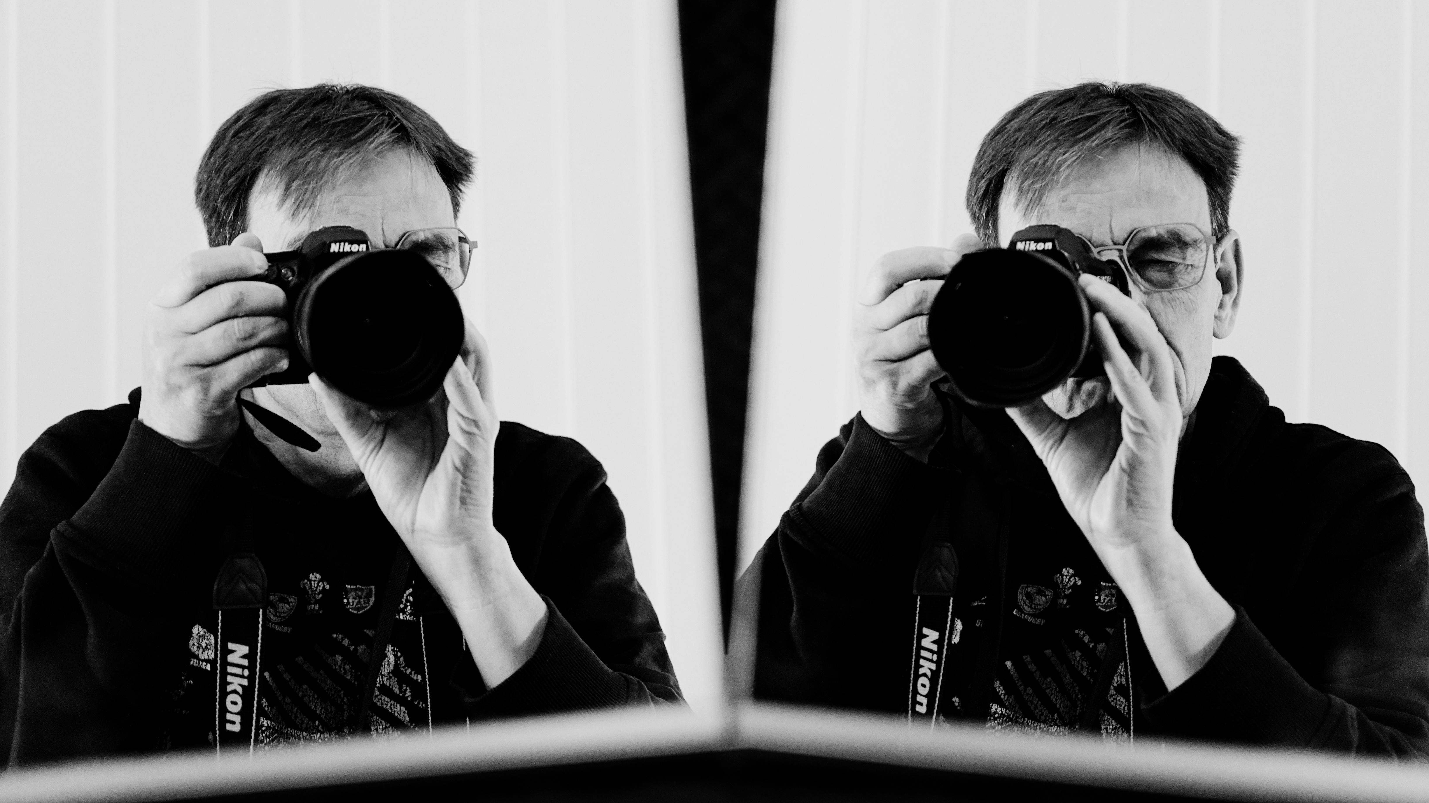 Man holding a Nikon camera close to his face, taking a photo, with his reflection visible in an angled mirror beside him. The image is in black and white, and the background features vertical lines. The man wears glasses and a dark sweatshirt, and the camera strap is visible around his neck.
###
Mann, der eine Nikon-Kamera nahe an seinem Gesicht hält und ein Foto macht, mit seinem Spiegelbild, das in einem abgewinkelten Spiegel neben ihm sichtbar ist. Das Bild ist schwarz-weiß und der Hintergrund verfügt über vertikale Linien. Der Mann trägt eine Brille und ein dunkles Sweatshirt, und der Kameragurt ist um seinen Hals sichtbar.