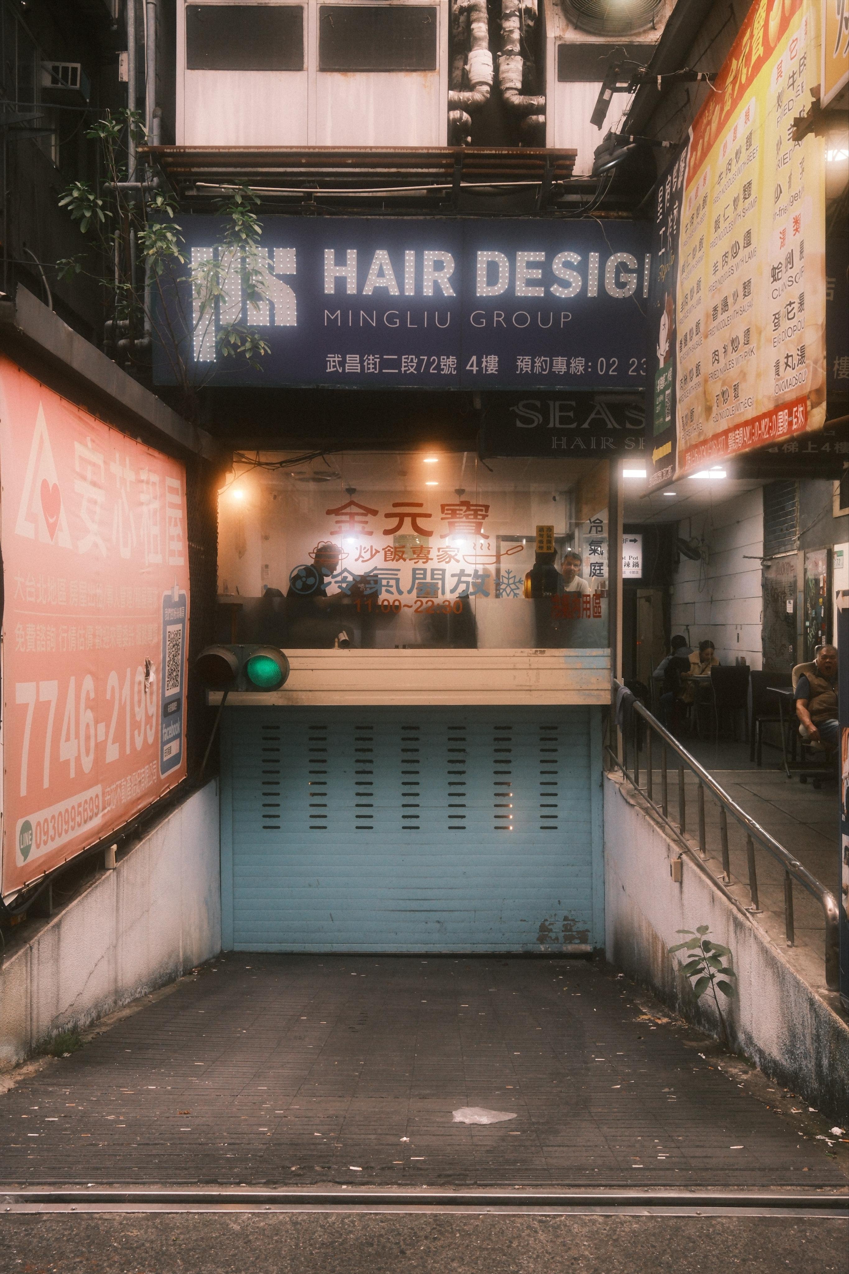 Narrow urban ramp leading down to an underground entrance, with a blue rolling shutter and a green traffic light at the bottom. Overhead and side walls are covered with Chinese and English signage, including a large “HAIR DESIGN / MINGLIU GROUP” sign, pipes, and hanging greenery; a lit interior corridor with a few seated people is visible on the right.