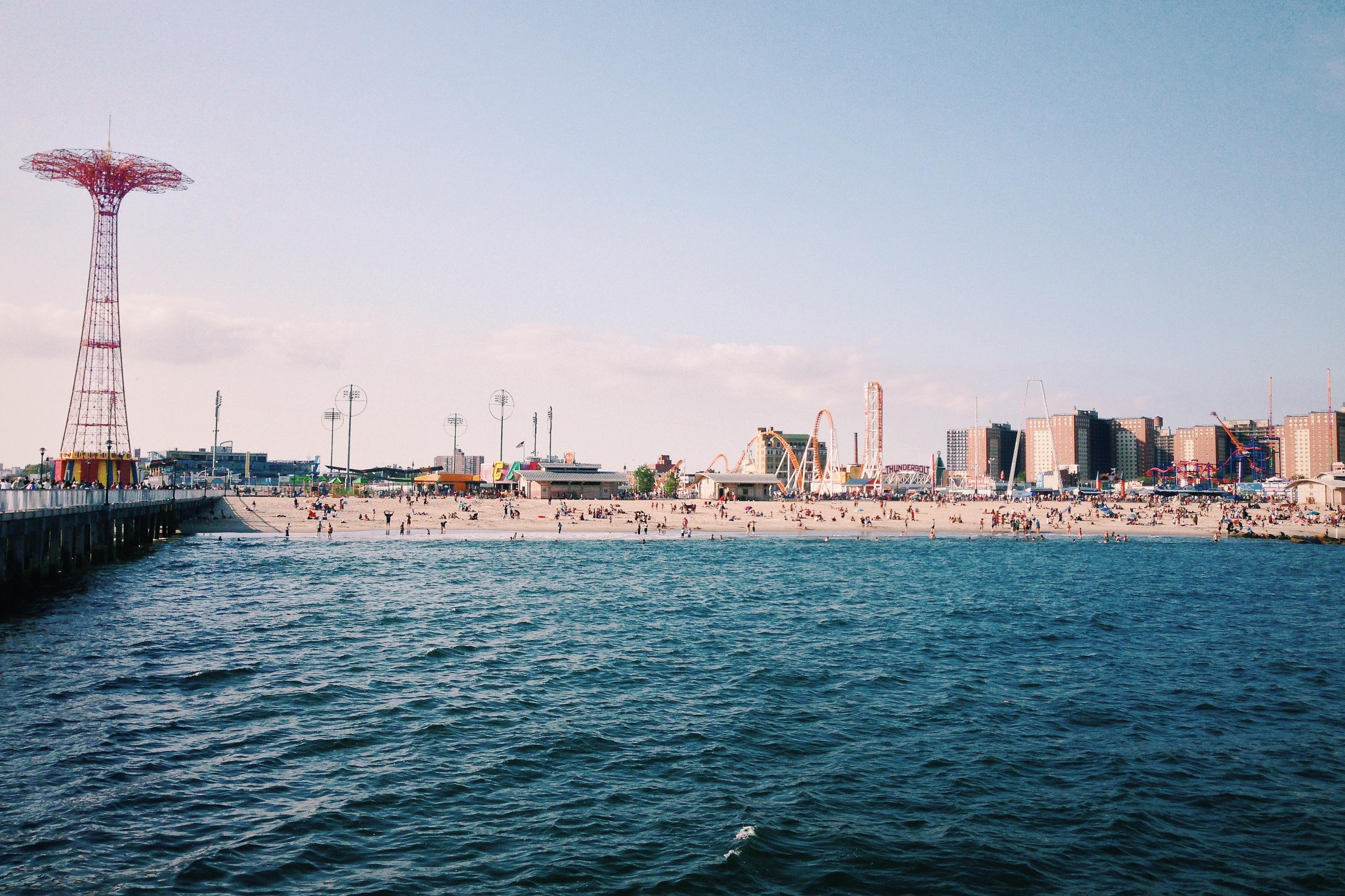 The image shows a beach and amusement park, Coney Island in Brooklyn, New York. It features a sandy beach with people, amusement park rides, and a tall red structure, which is the Parachute Jump. There's water in the foreground and buildings in the background.