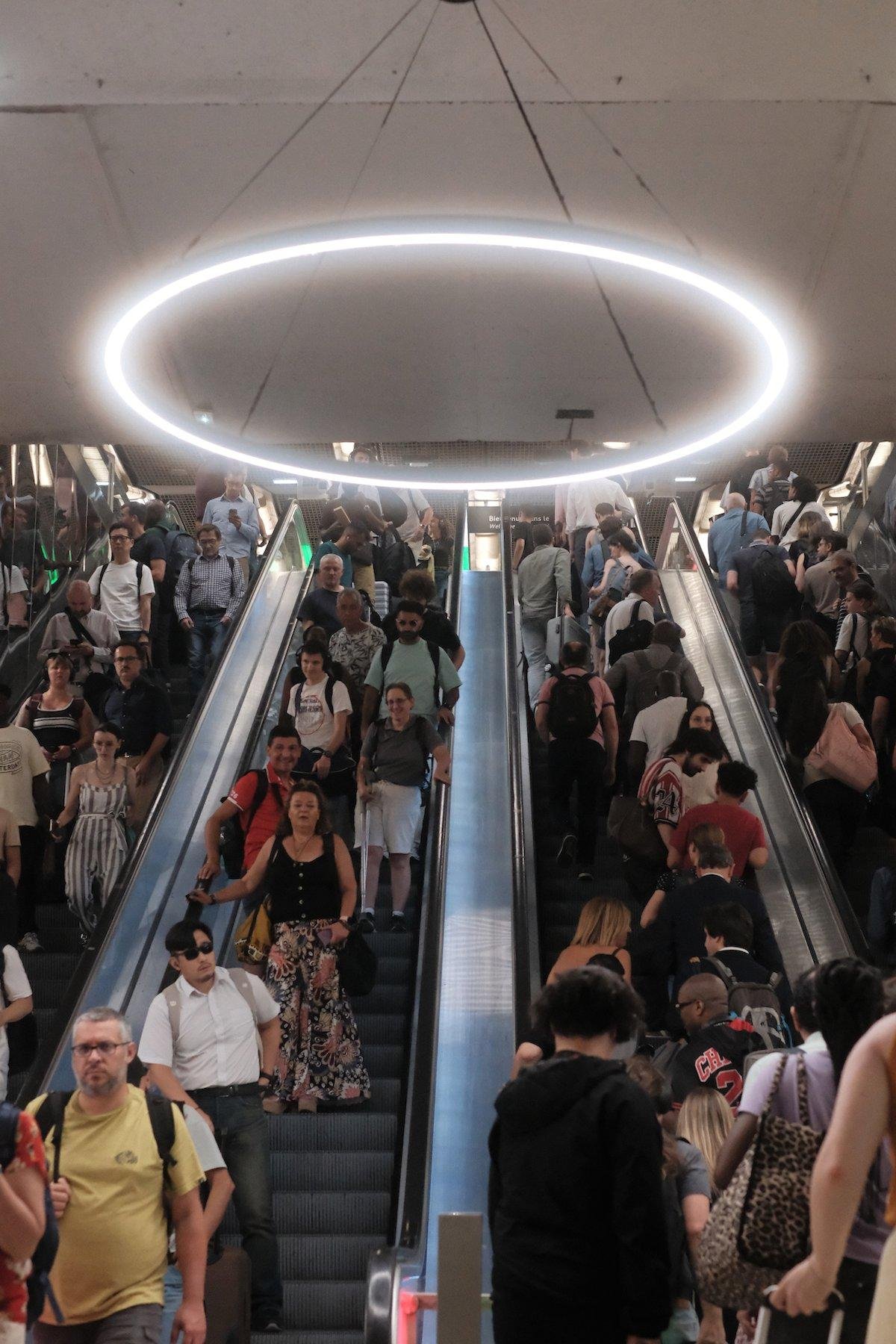 Photo verticale. 4 escalators, 2 dans chaques sens, plein de gens et au plafond, un énorme cercle lumineux éclaire le passage