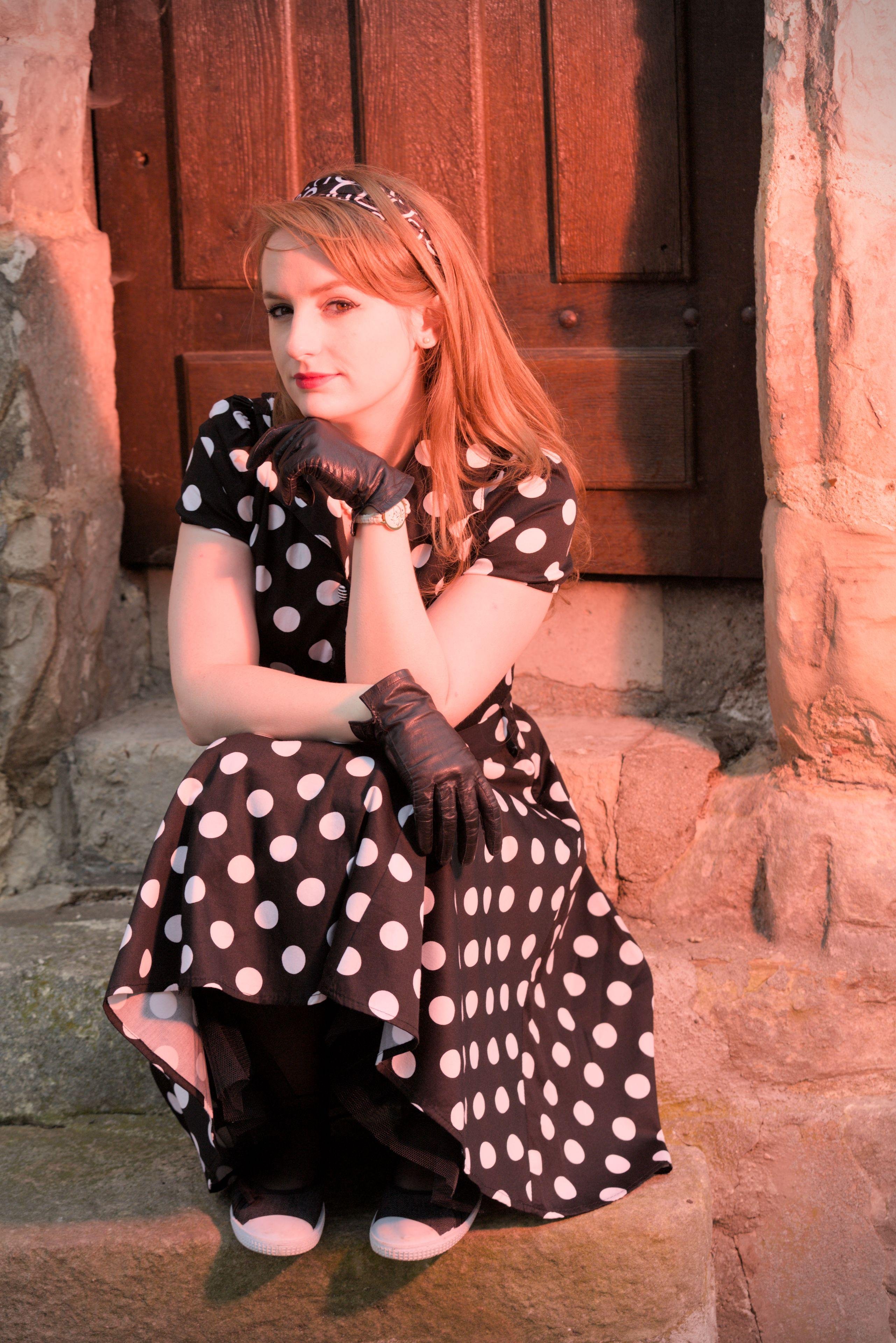 Une jeune femme rousse dans une robe noire à gros pois blanc, avec des gants noirs en cuir et un serre tête, assise sur des marches en pierre d’un petit escalier de 3 marches menant à une porte. Sa main gauche est juste en dessous de son menton, sa tête est tournée vers sa droite et ses yeux regardent la caméra