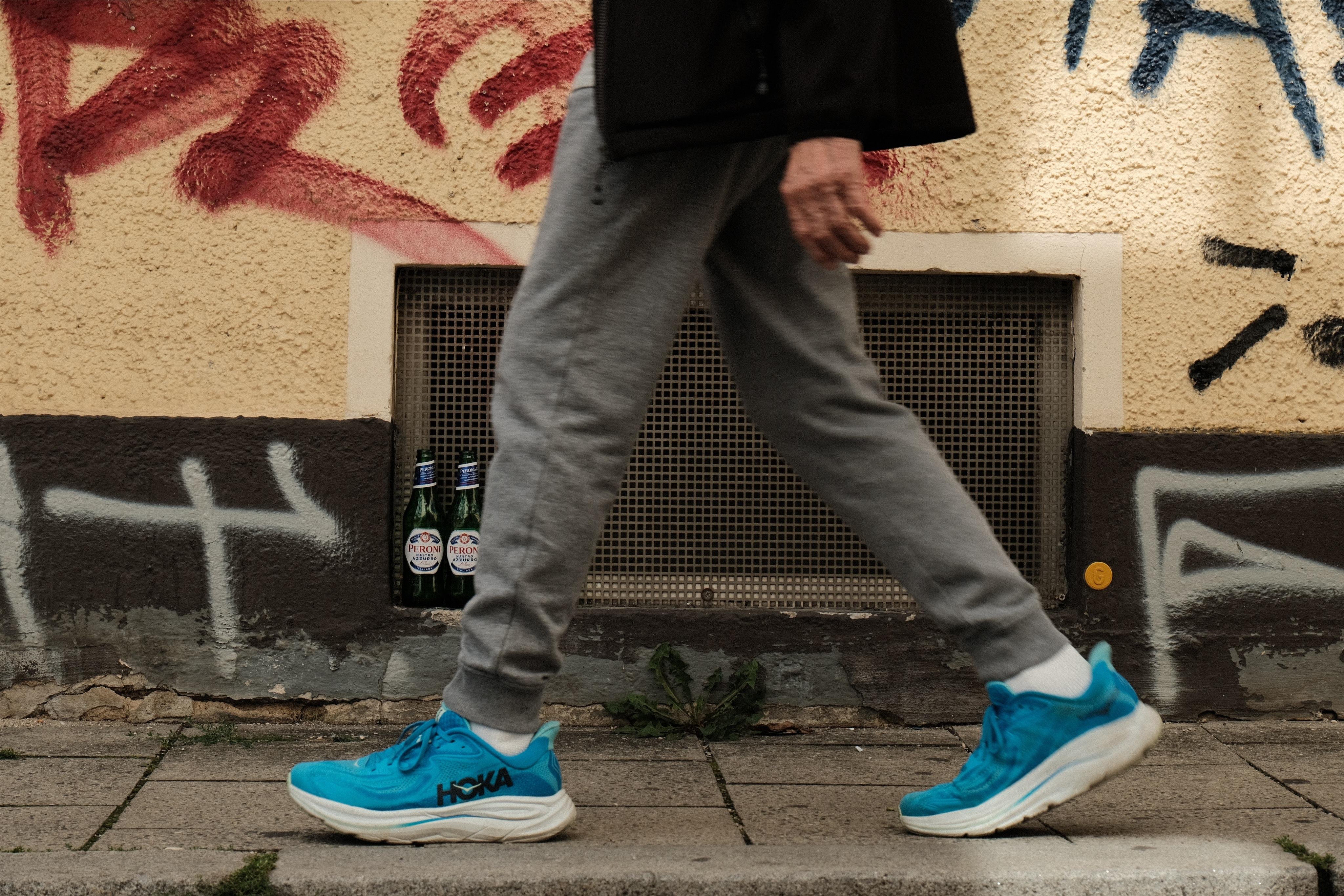 An old man with new blue sneakers walking oat a yellow wall with graffiti. In the cellar window in the background someone put empty beer bottles.
You can only see the legs of the walking man. Shot from the same height as the shoes.