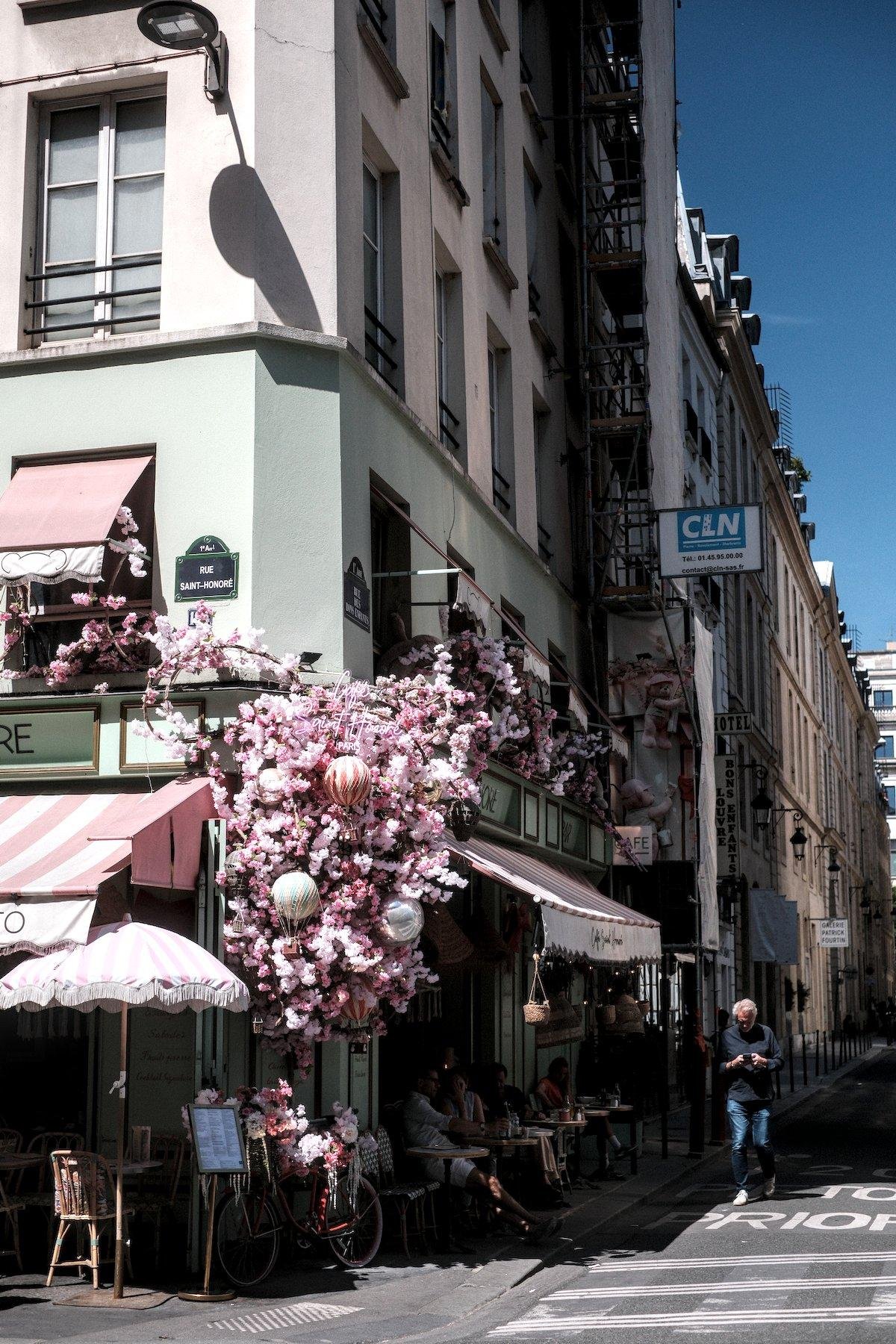 Photo verticale. Angle de rue parisienne occupé par un café qui a mi une énorme décoration en fleurs rose pile à l’angle, contrastant avec les murs peints en vert sauge