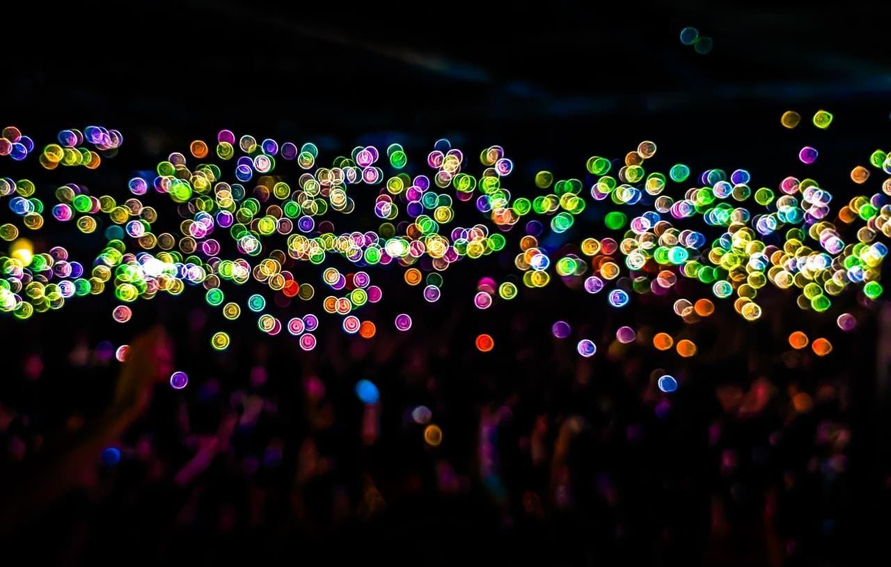 Colorful bokeh lights create an abstract pattern against a dark background. The lights vary in color, including pink, yellow, green, blue, and purple, and appear as scattered circles of different sizes. The overall effect is vibrant and festive.