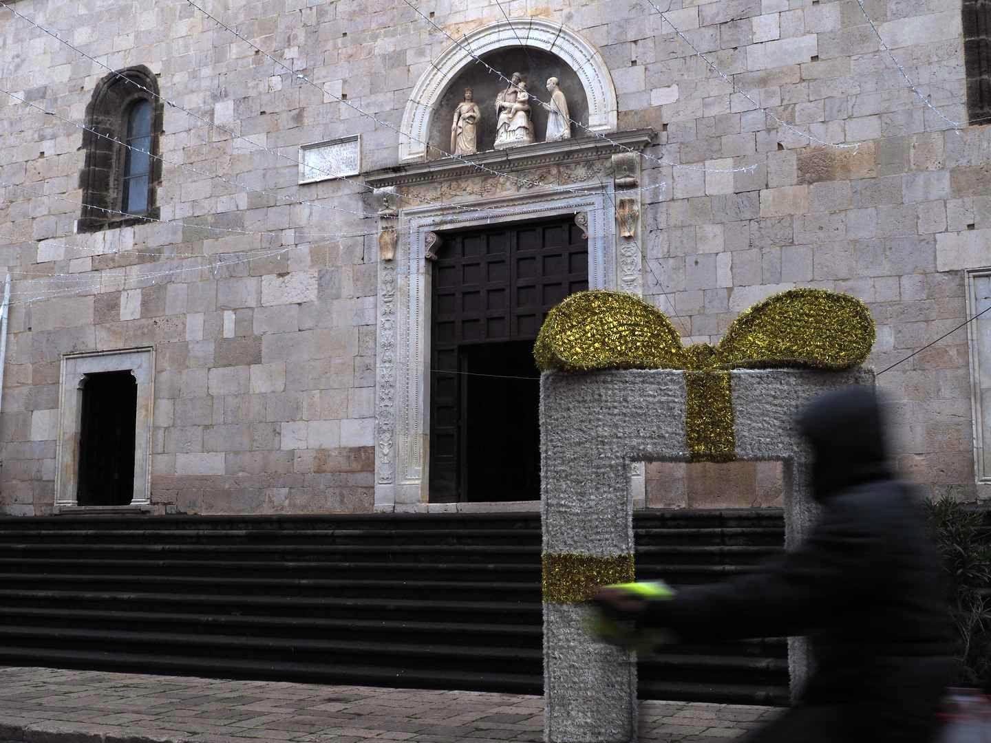 Facciata di una chiesa, con una scalinata e un trittico di statue sull'ingresso principale, all'interno di un arco. In basso a destra, un soggetto sfocato in bicicletta sfreccia davanti a una decorazione natalizia.