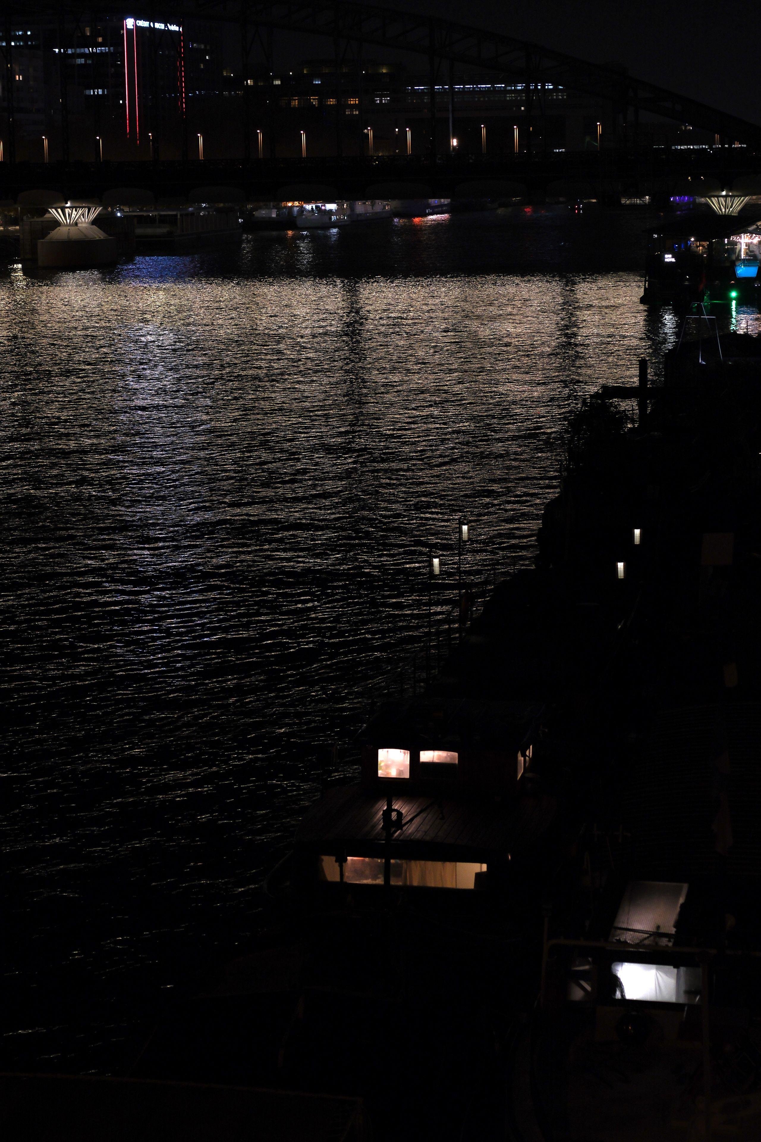 Photo format portrait de nuit, montrant la Seine et des péniches sur la droite, avec de la lumière qui en émane. Au fond, un pont avec des lampadaires et les pieds du pont éclairés