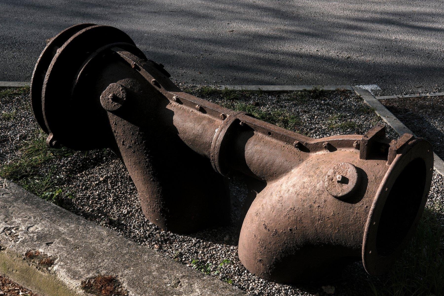 Photo horizontale. Une structure en métal couleur rouille, 2 gros tuyaux entrent dans le sol, un tuyau horizontal les relie, et ça se fini en disque de chaque côté, le tout éclairé par une lumière de soleil couchant