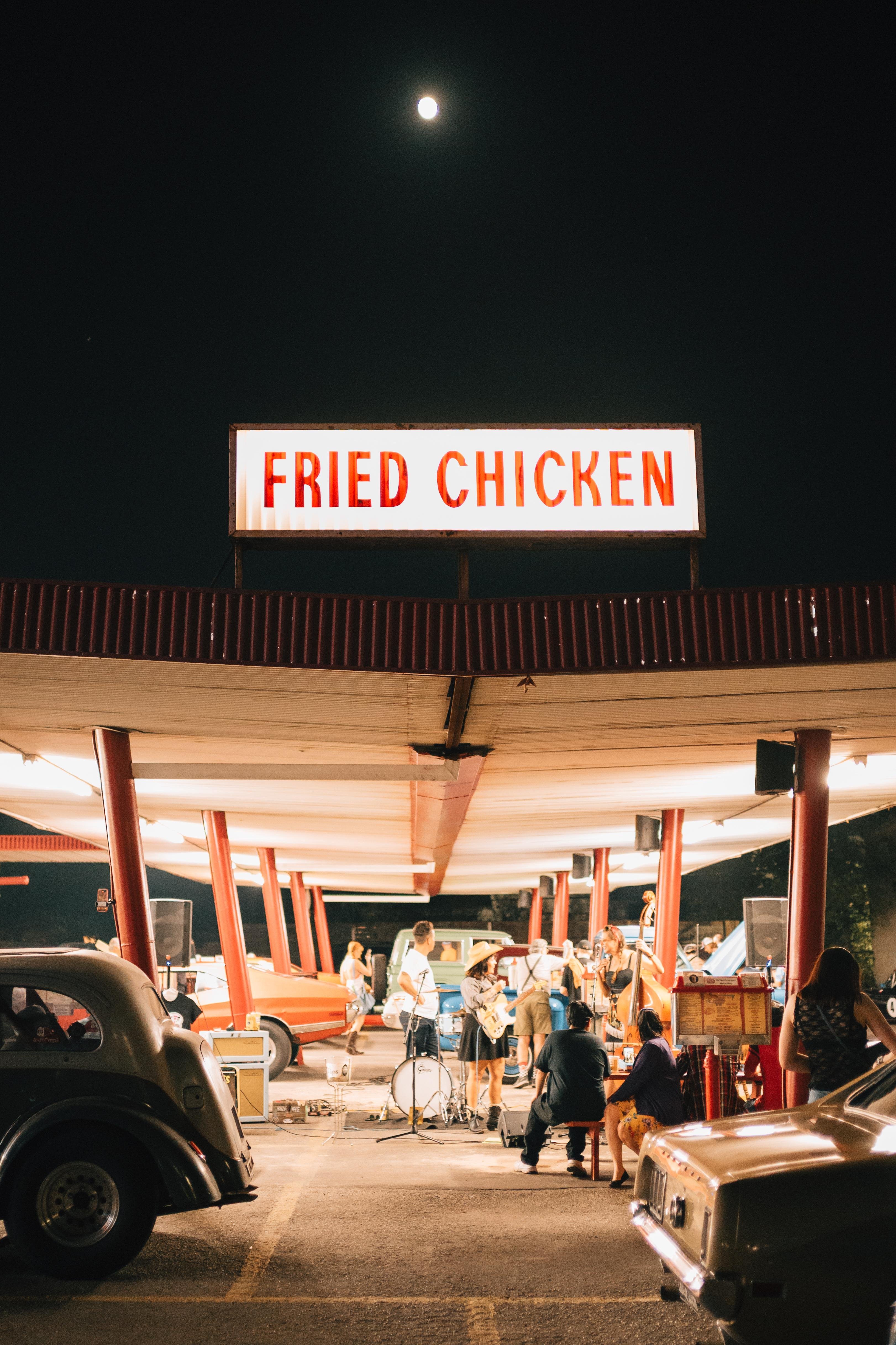 Moonlit scene at a drive-in with a large "Fried Chicken" sign. Vintage cars are parked under a canopy with red poles. A band is performing with various instruments, while people are gathered around. The night sky is visible in the background.